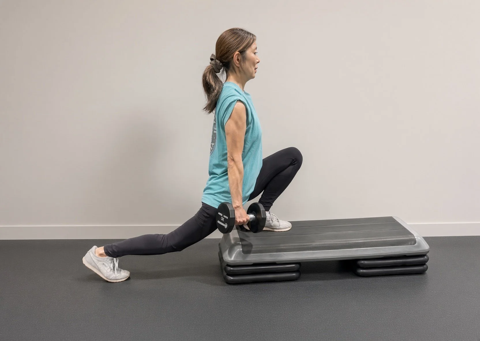 A woman is doing a lunge exercise with dumbbells, stepping onto a riser at the gym.