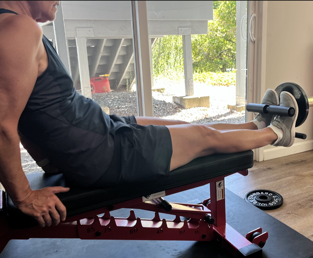 A man exercising on a workout bench with a barbell on his legs to perform leg raise exercise, near a sliding glass door looking outside.