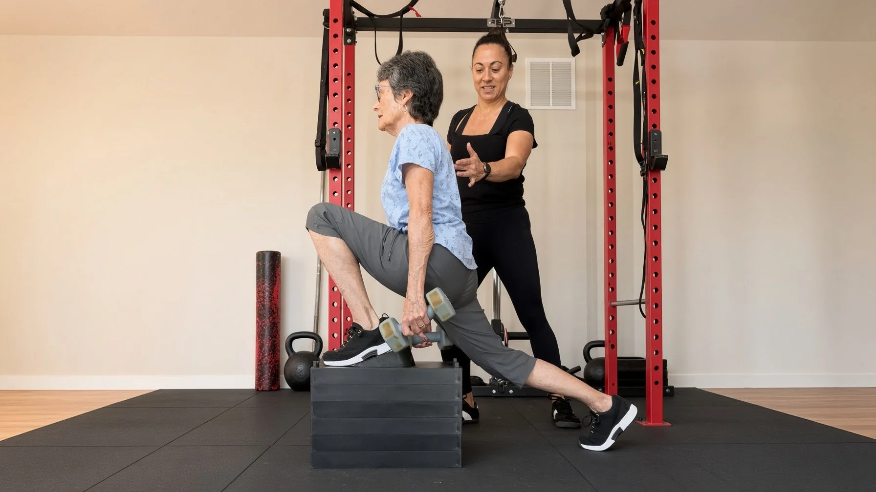 An elderly woman in workout clothes performs a step-up exercise with dumbbells under the supervision of a trainer in a home gym.