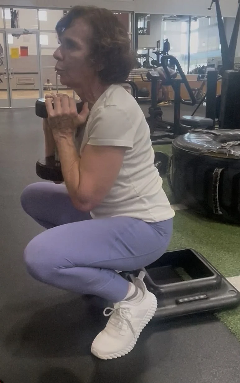 An elderly woman in workout clothes performing a squat with a dumbbell in a gym.