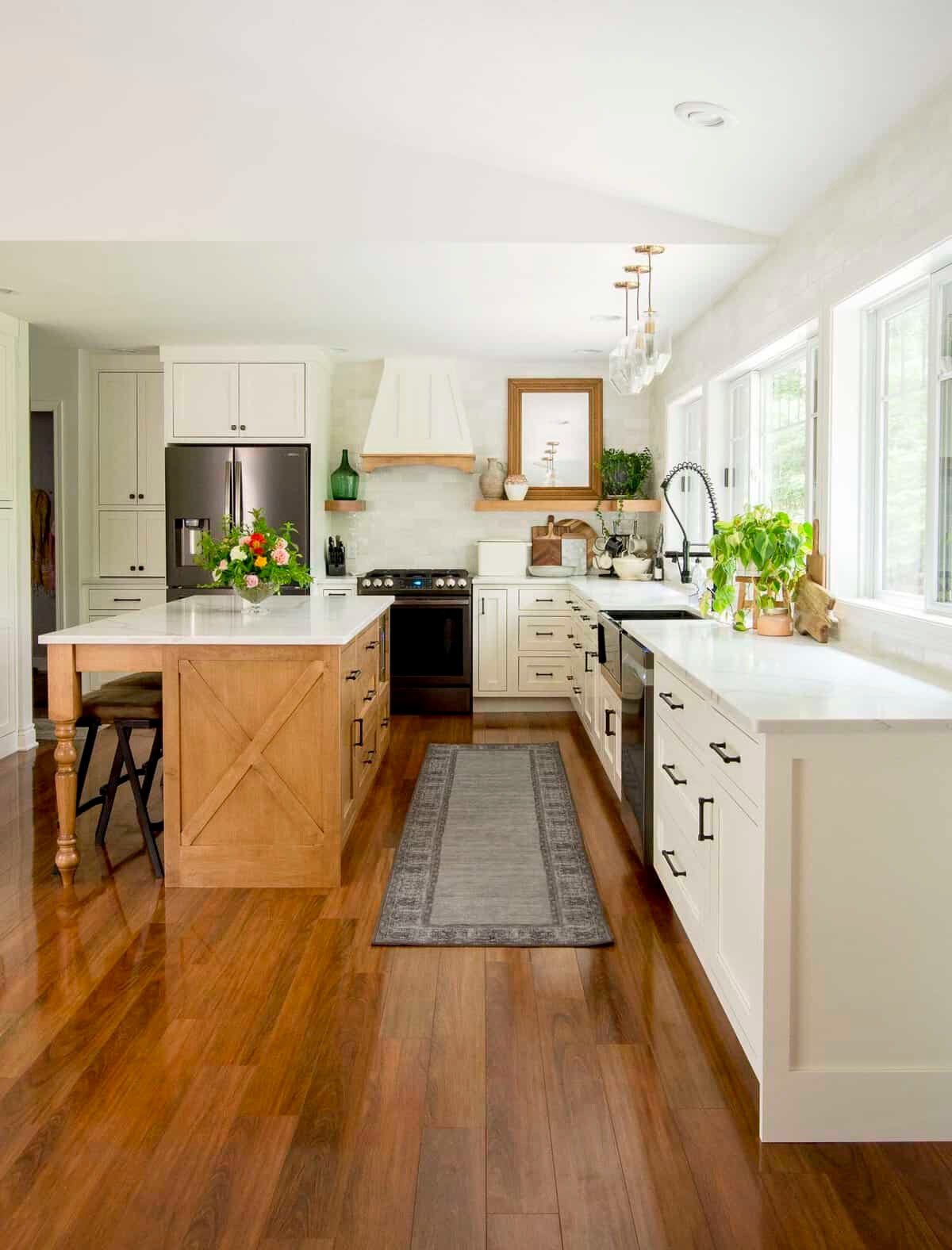 Bright kitchen with white cabinets, wooden island, and hardwood flooring. Decor includes plants, a flower arrangement, framed art, and kitchen essentials near a large window.