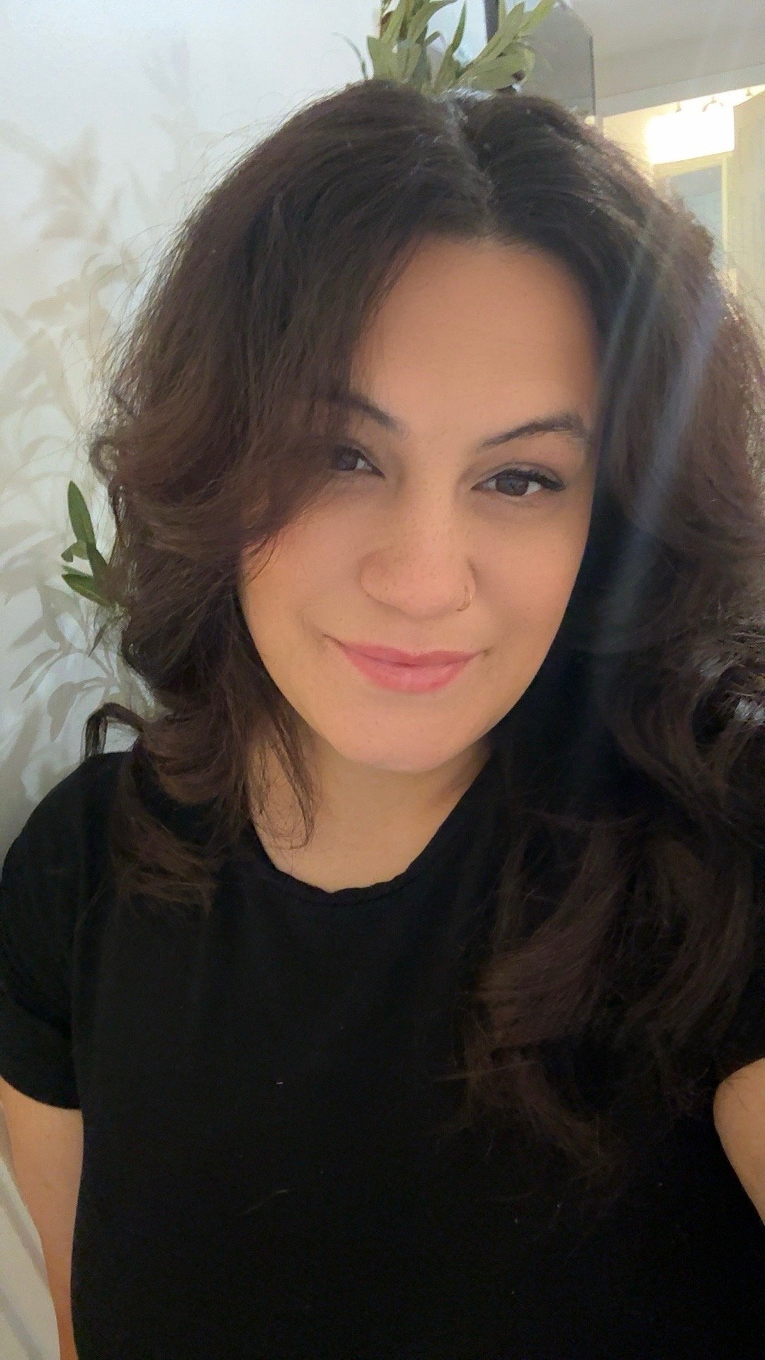 A woman with dark brown, wavy hair, wearing a black shirt, smiling softly, standing in front of a light-colored wall with a plant and a bright window or light source in the background.