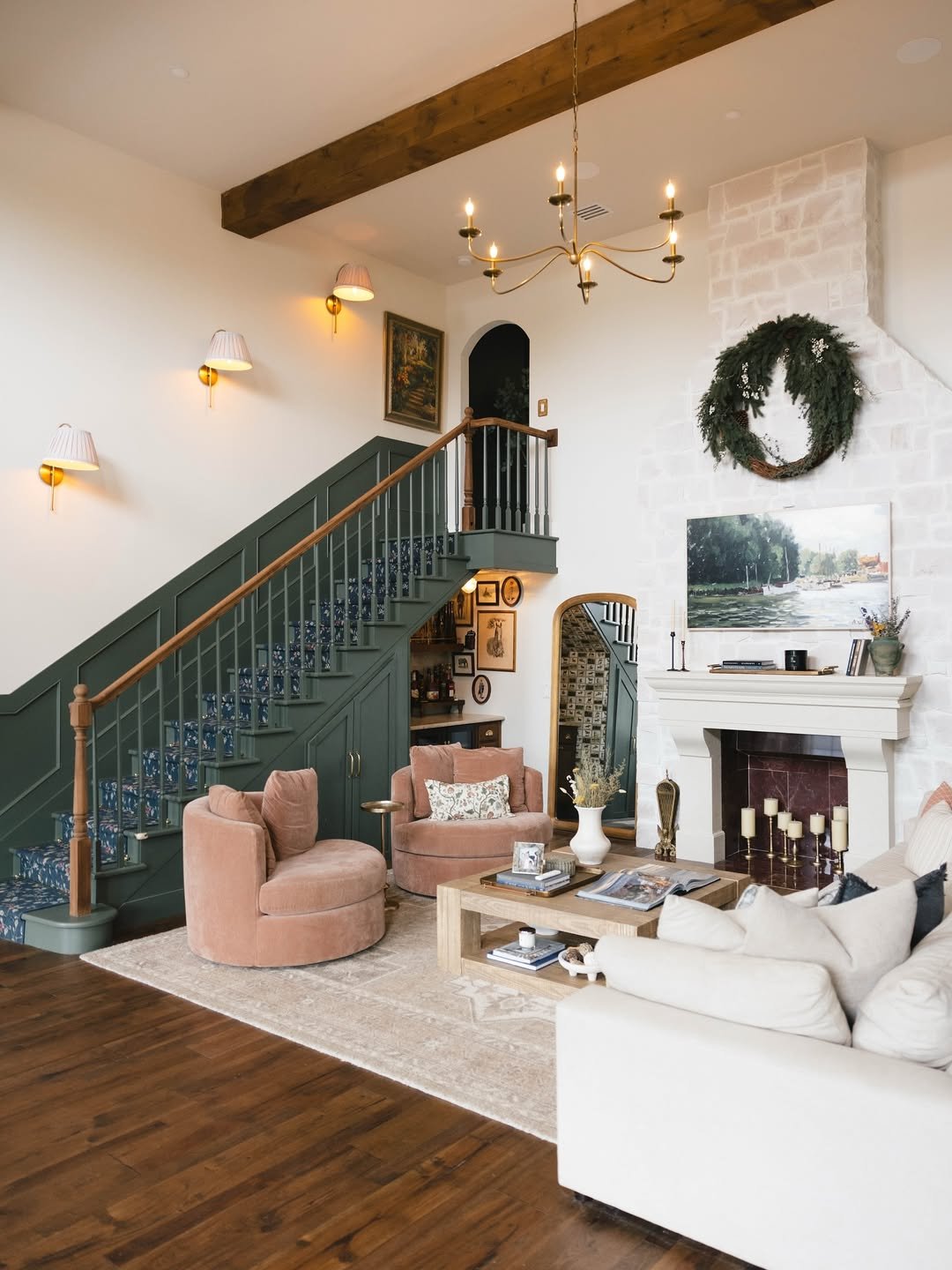 Living room with fireplace, beige sofa, pink velvet chairs, wooden coffee table, dark green staircase with floral carpet, wall art, wall sconces, chandelier, wreath, and a landscape painting above the fireplace.