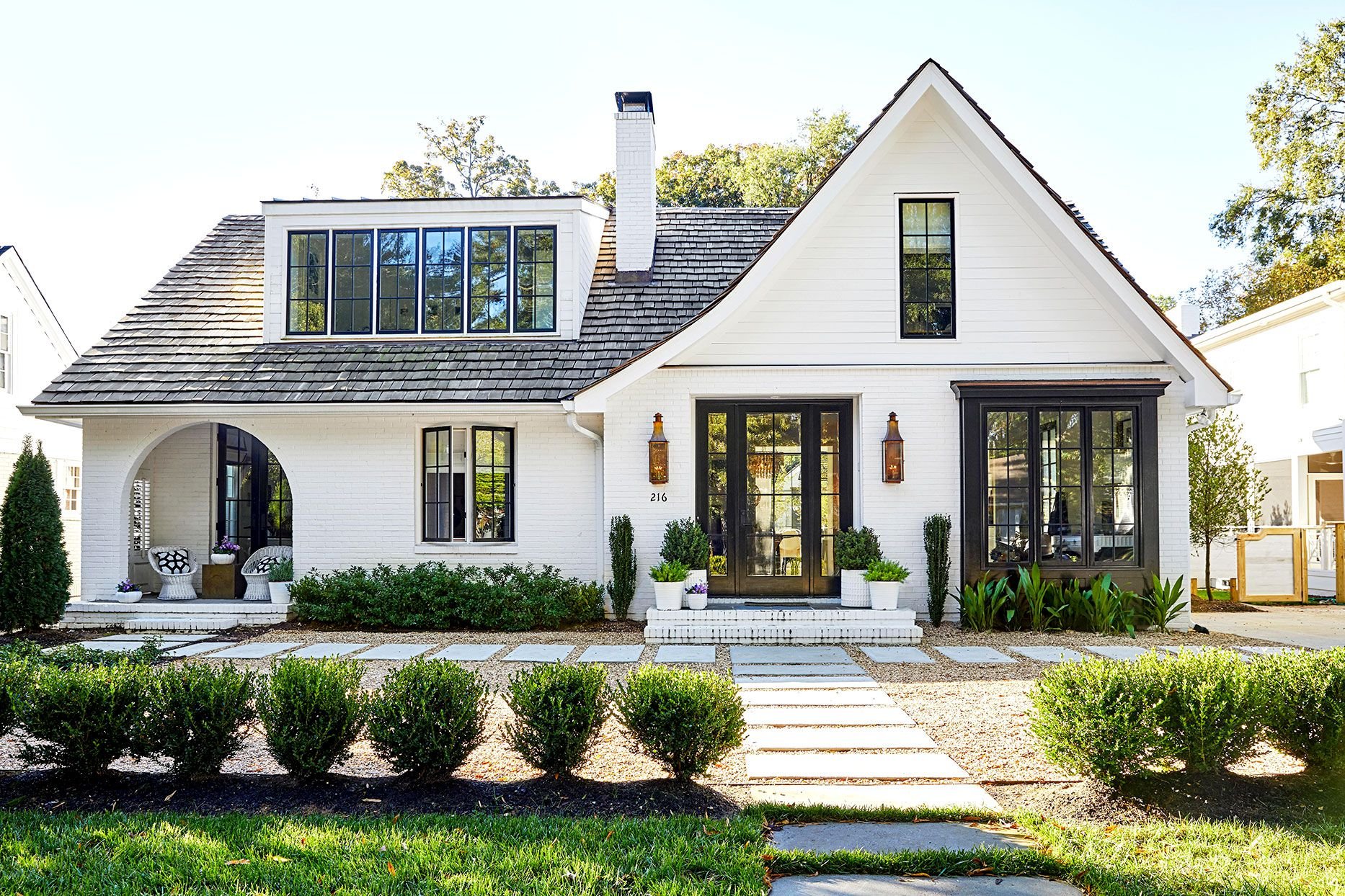 Front view of a modern white house with large black-framed windows, a gabled roof, a brick chimney, and landscaped front yard with bushes and a pathway.