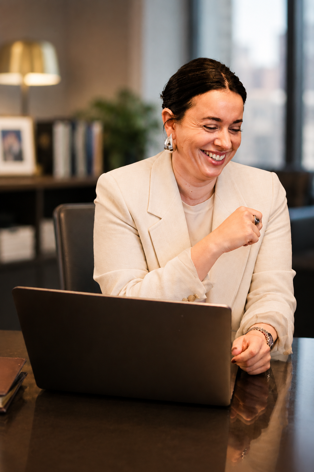 A woman in a beige blazer sitting at a dark wooden table, smiling while looking down at her laptop, in a modern office setting.
