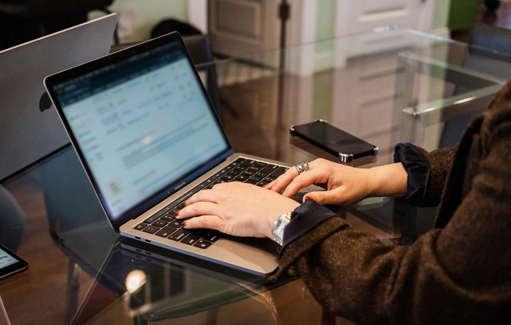 Person using a laptop with a blurred screen, sitting at a glass table, with a smartphone nearby.