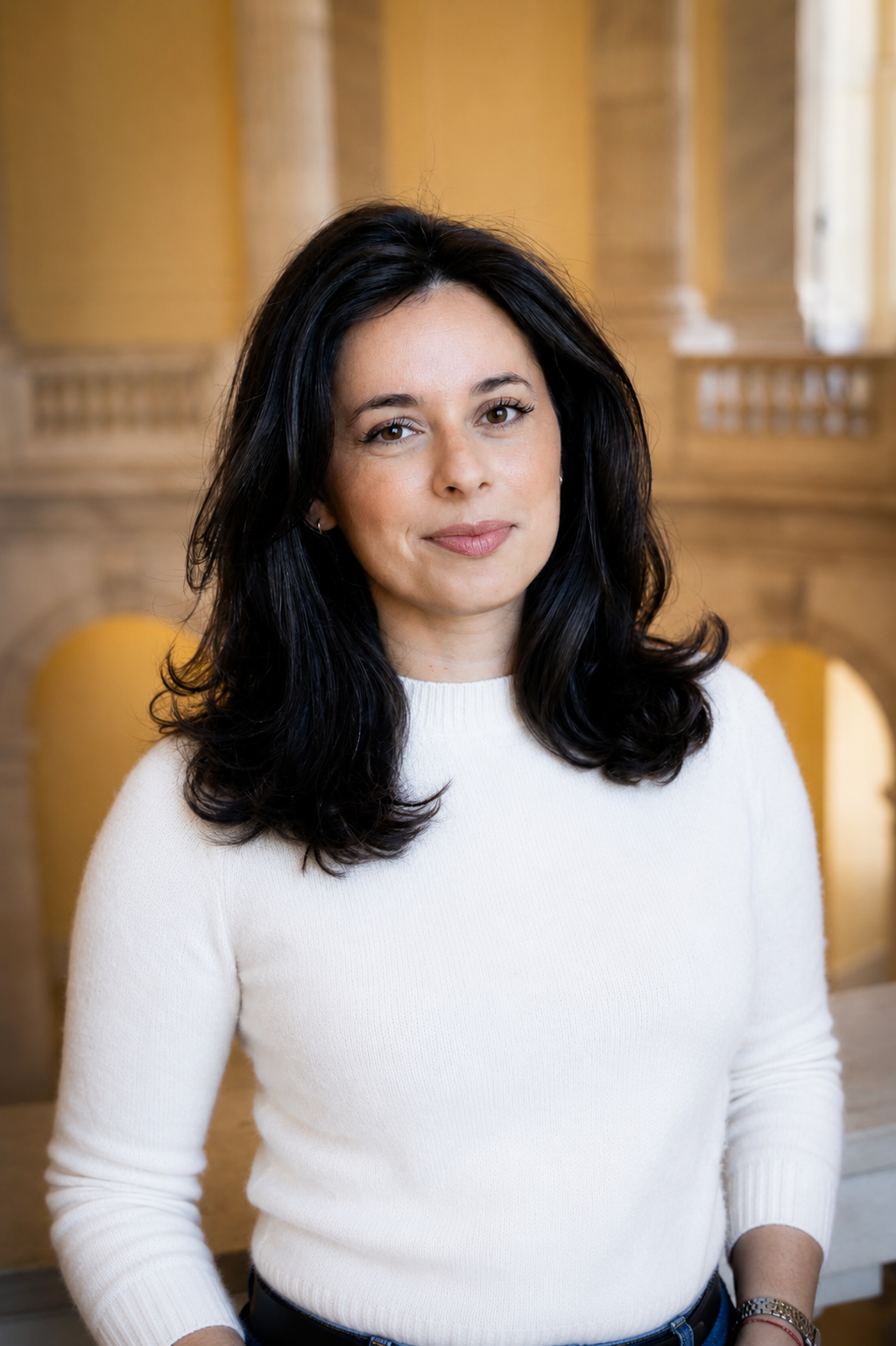 A woman with dark hair and a white sweater standing in an ornate, warmly lit interior with wooden pillars and arches.