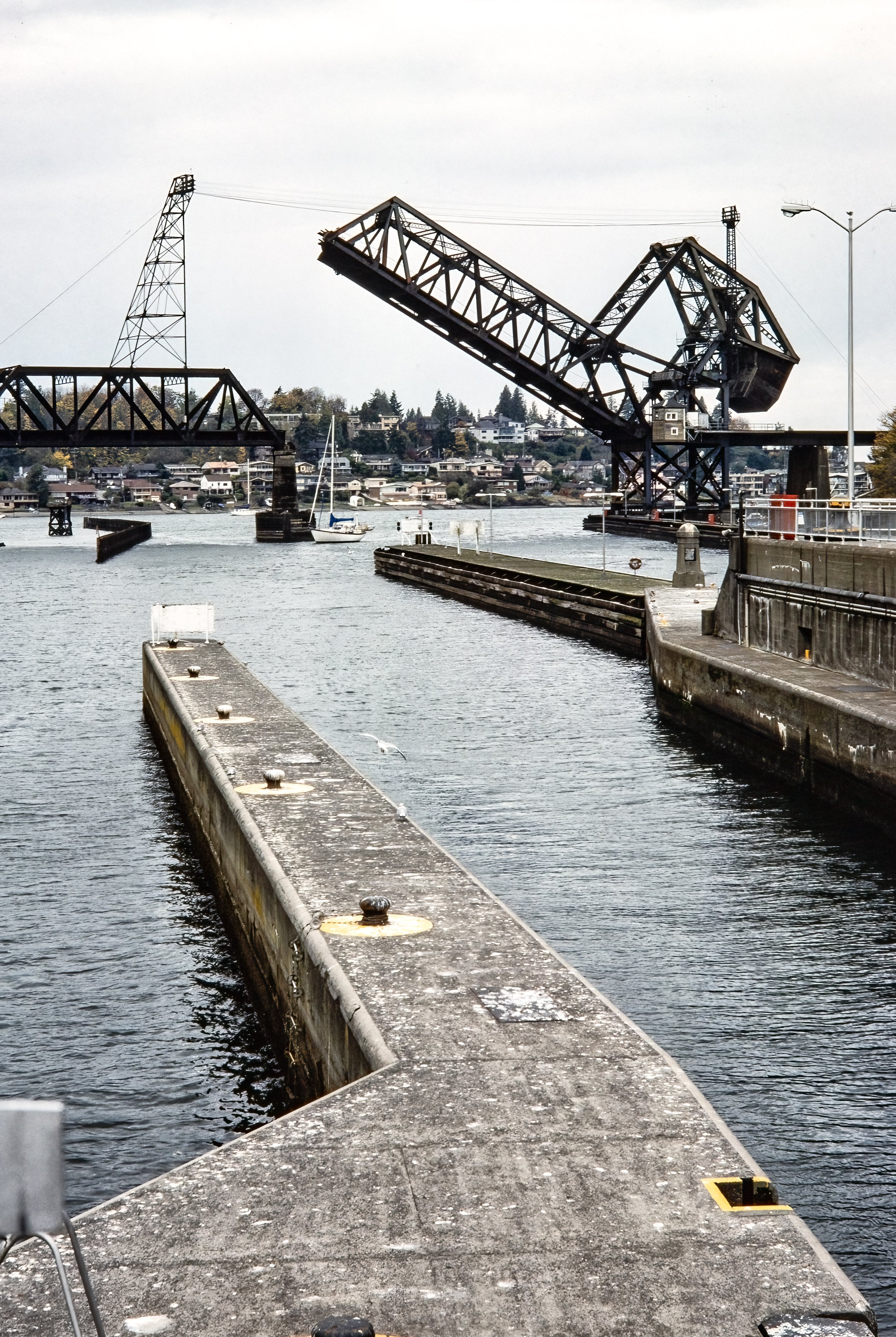 "Lifted Passage – Hiram Chittenden Locks and Railway Bridge" - A sailboat moves through the Hiram Chittenden Locks as the adjacent railway bridge rises, showcasing the seamless dance of industry and waterway in Seattle WA.