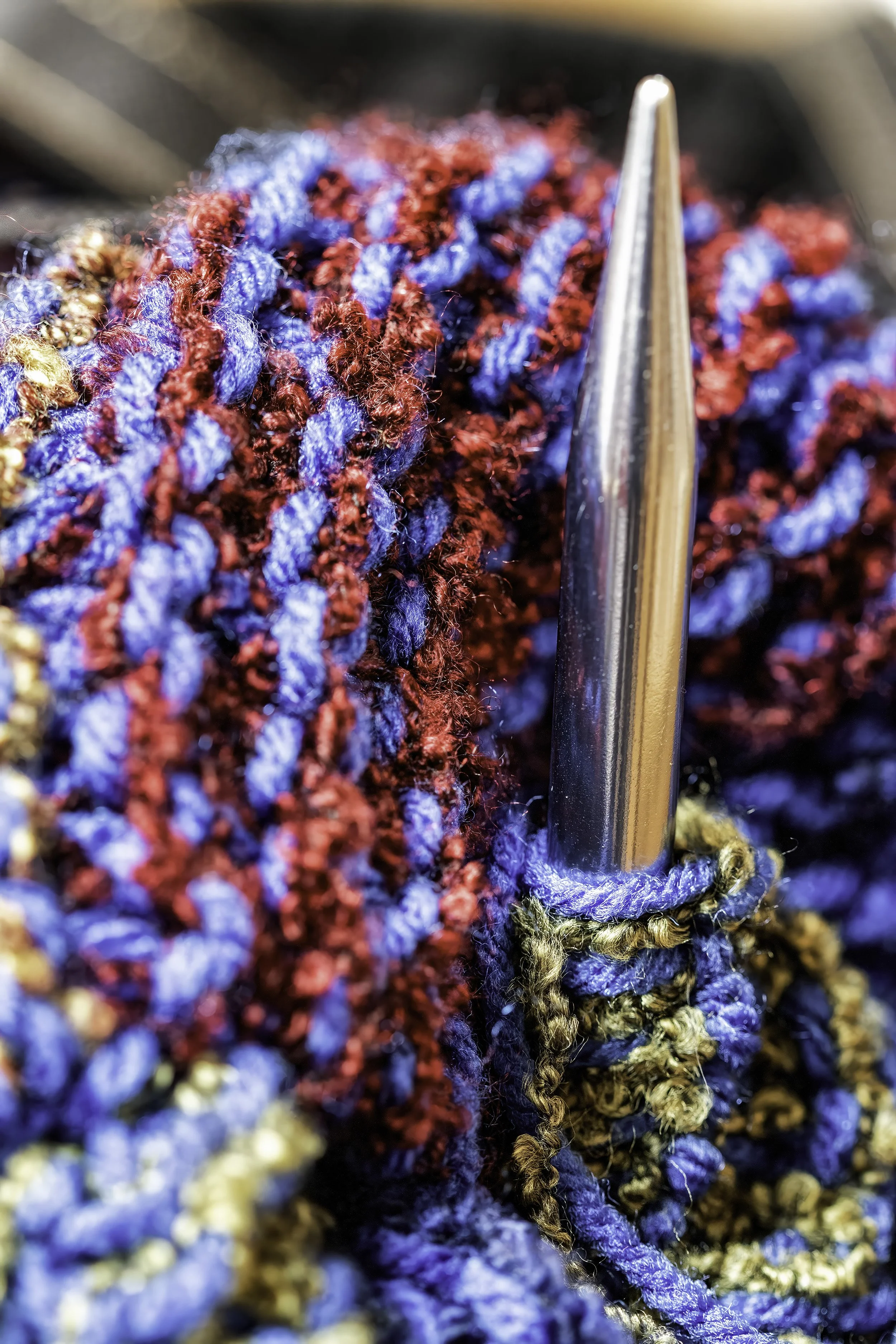 "Needle & Hue – A Symphony in Yarn"-A richly textured macro image showcasing a metal knitting needle nestled in vibrant blue, red, and olive yarn. The close-up captures the tactile beauty and rhythm of handcraft.