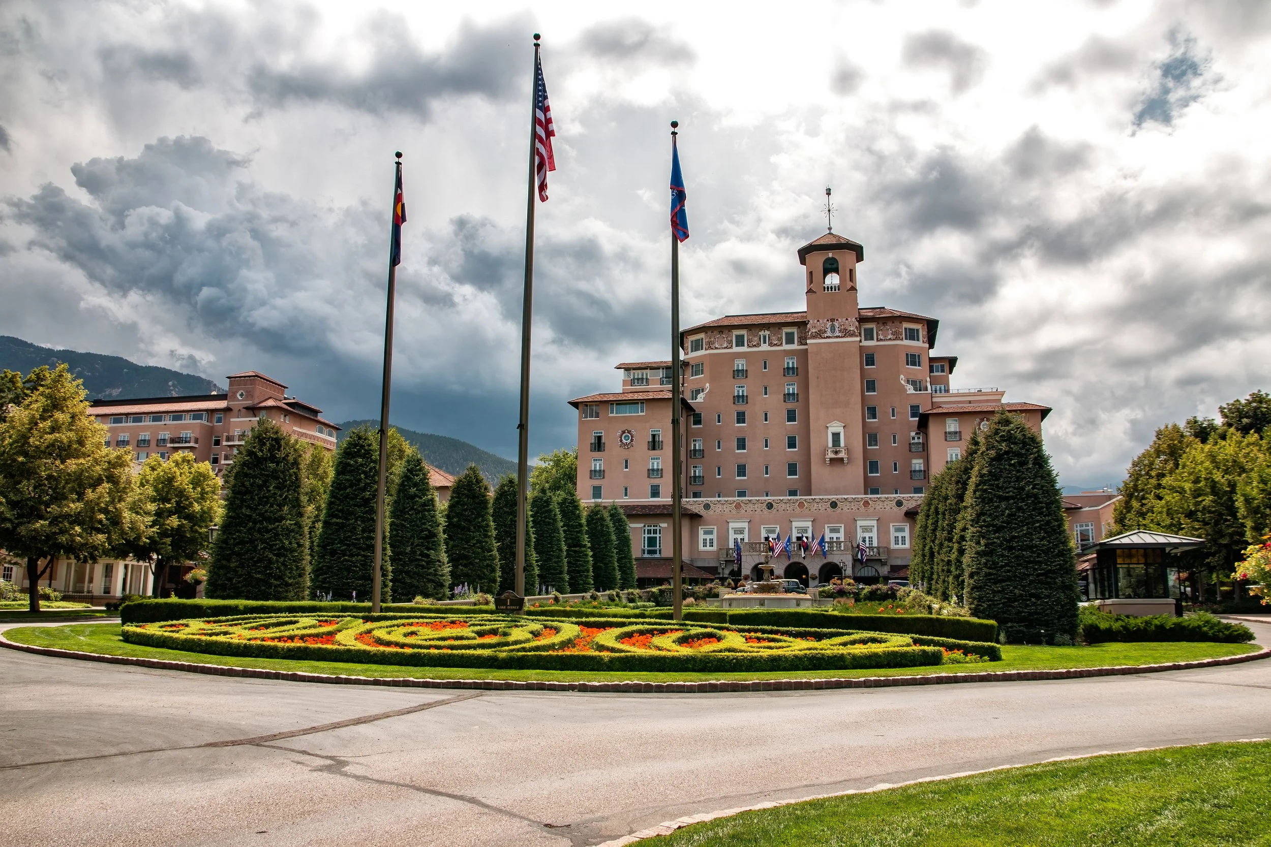 "Entrance to The Broadmoor" - The Broadmoor's grand entrance, framed by lush gardens and flags, set against the dramatic Rocky Mountains near Colorado Springs Colorado.