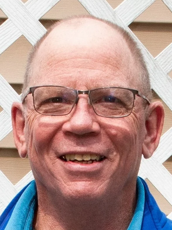 A middle-aged man with short hair, glasses, and a big smile, wearing a blue jacket, standing in front of a wooden lattice background.