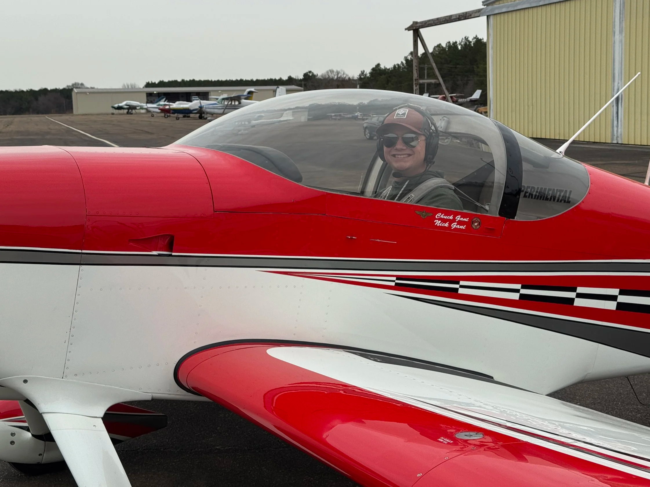 A smiling man in sunglasses and a cap sitting inside a red and white experimental aircraft at an airport, with a row of small airplanes and hangars in the background.