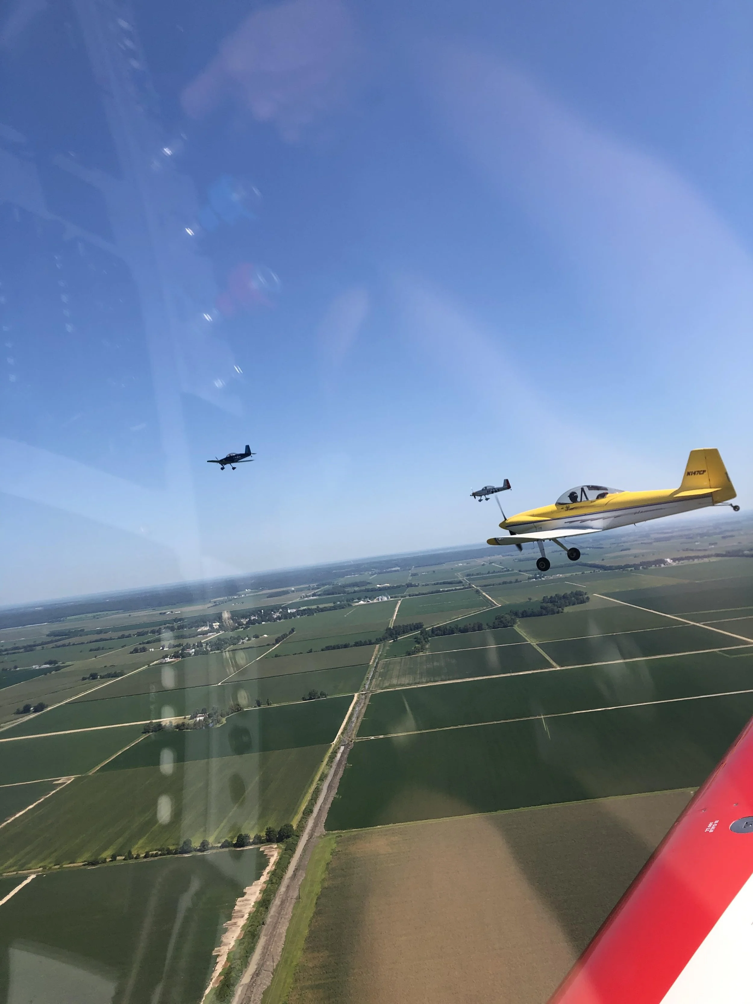 View from an airplane window showing three small planes flying over farmland and fields, with a blue sky above.