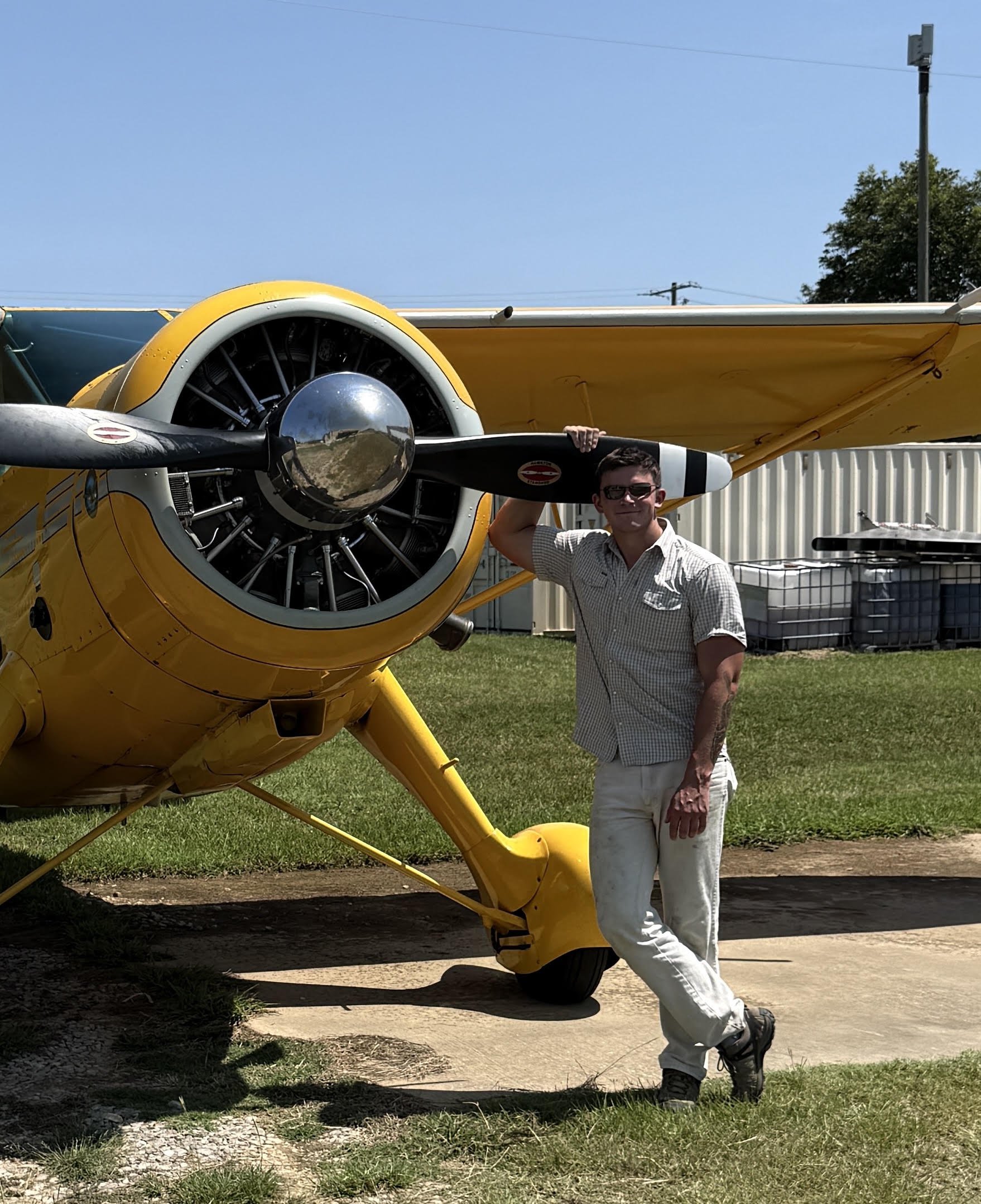 A man with sunglasses and tattoos posing next to a yellow and black aircraft on a grassy area with a fence and trees in the background.