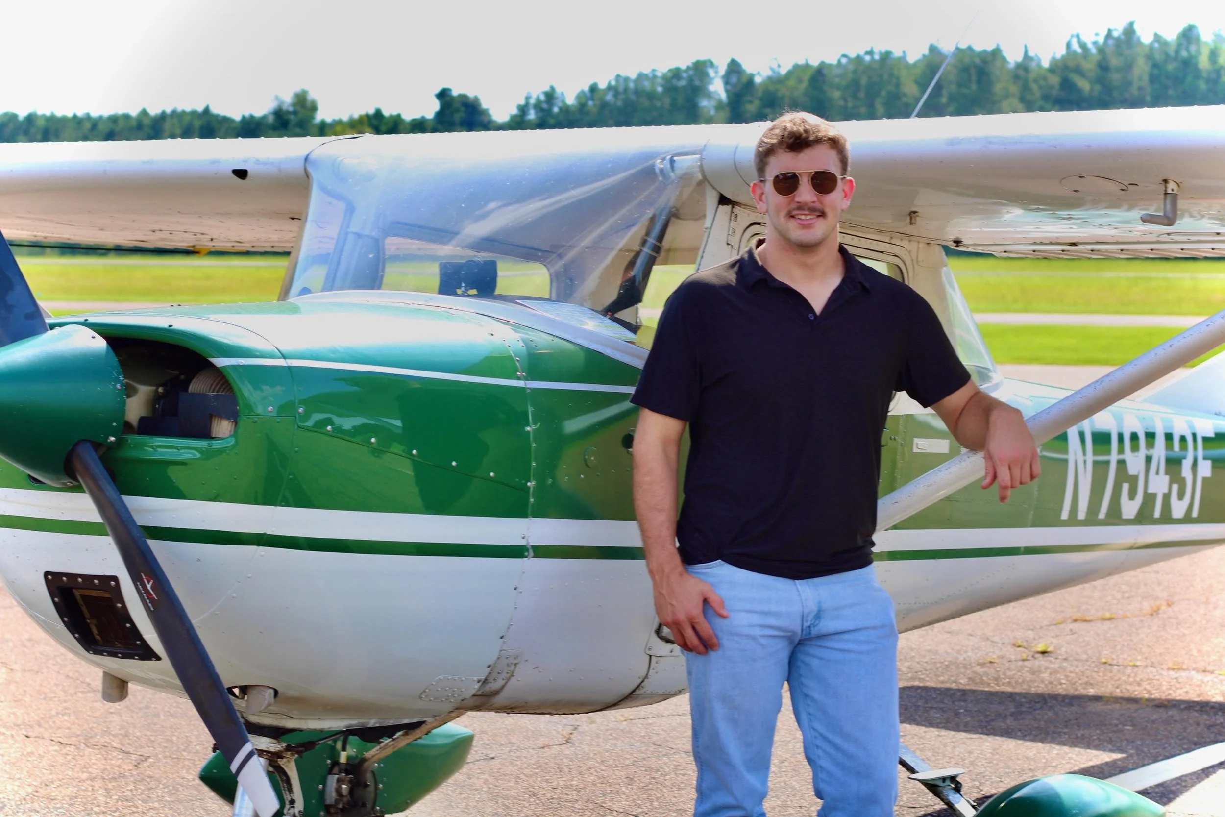 A man with sunglasses, wearing a black shirt and light blue jeans, standing next to a small green and white airplane outdoors, with a background of trees and clear sky.