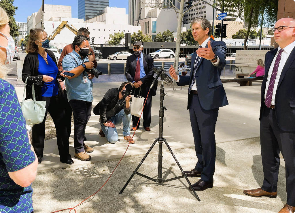 A man in a navy suit and tie speaking at a microphone outdoors to an audience, with several people listening and taking photos in an urban setting with buildings and construction in the background.