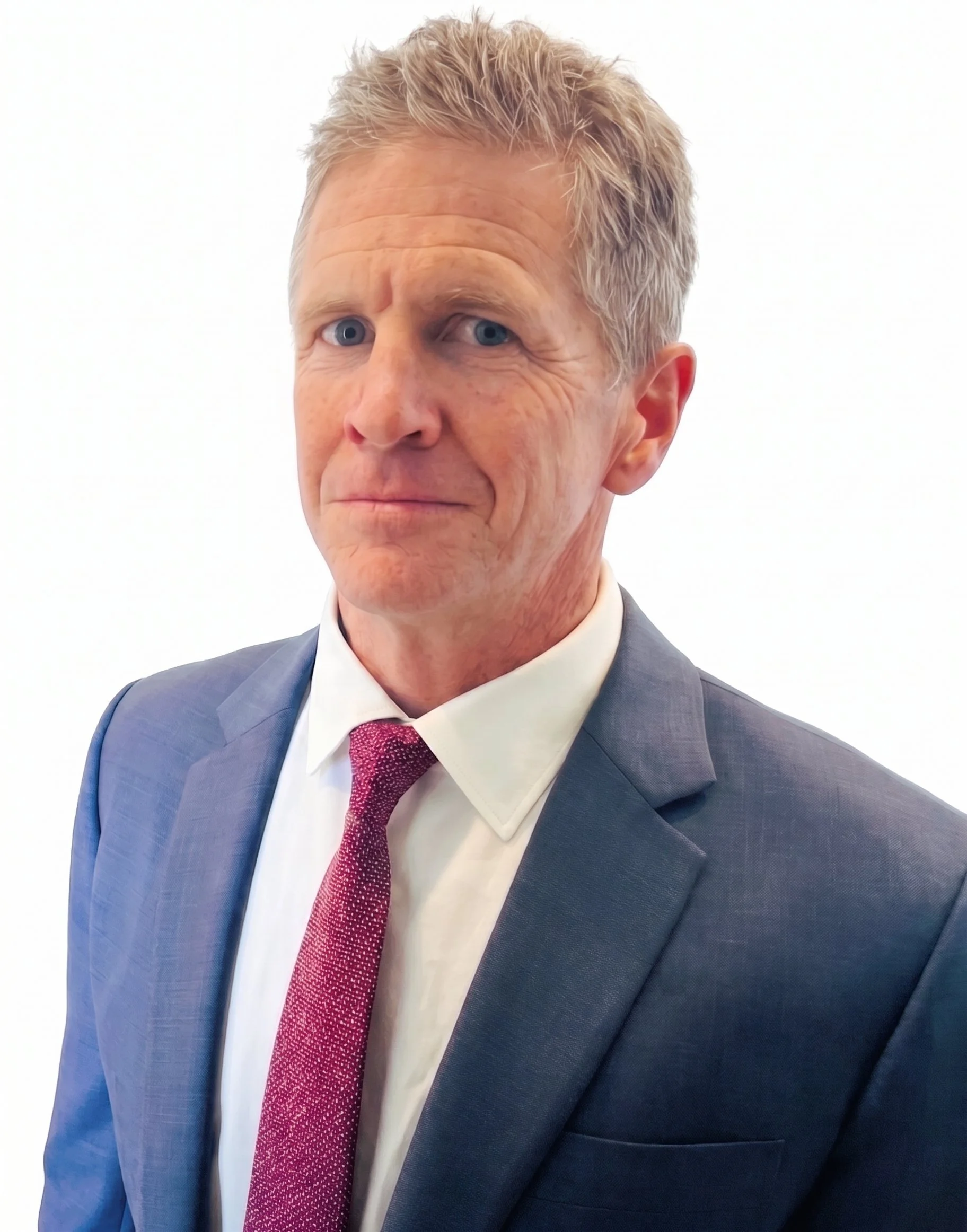 A middle-aged man with short, gray hair and blue eyes wearing a blue suit, white shirt, and red patterned tie against a plain white background.