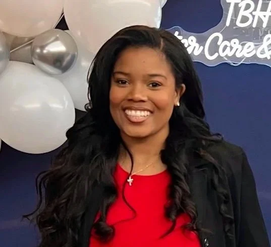 A smiling woman with long dark hair, wearing a red top and a black jacket, standing in front of a blue backdrop with white balloons and a sign.