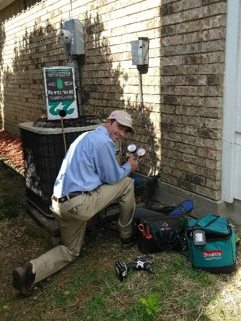 A man working on an air conditioning unit outside a brick building, kneeling with tools and equipment nearby.