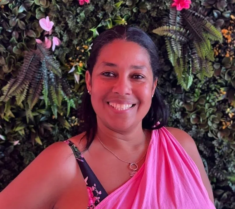 A smiling woman wearing a pink saree with a black floral blouse, standing in front of a lush green wall with pink and white flowers.