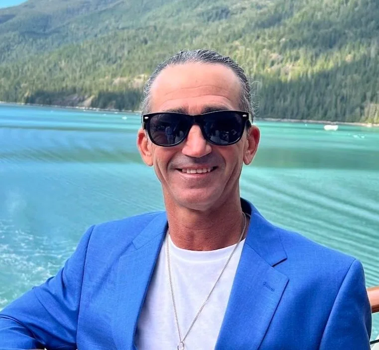 A smiling man wearing black sunglasses, a blue blazer, and a white shirt, posing outdoors with a lake and forested hills in the background.
