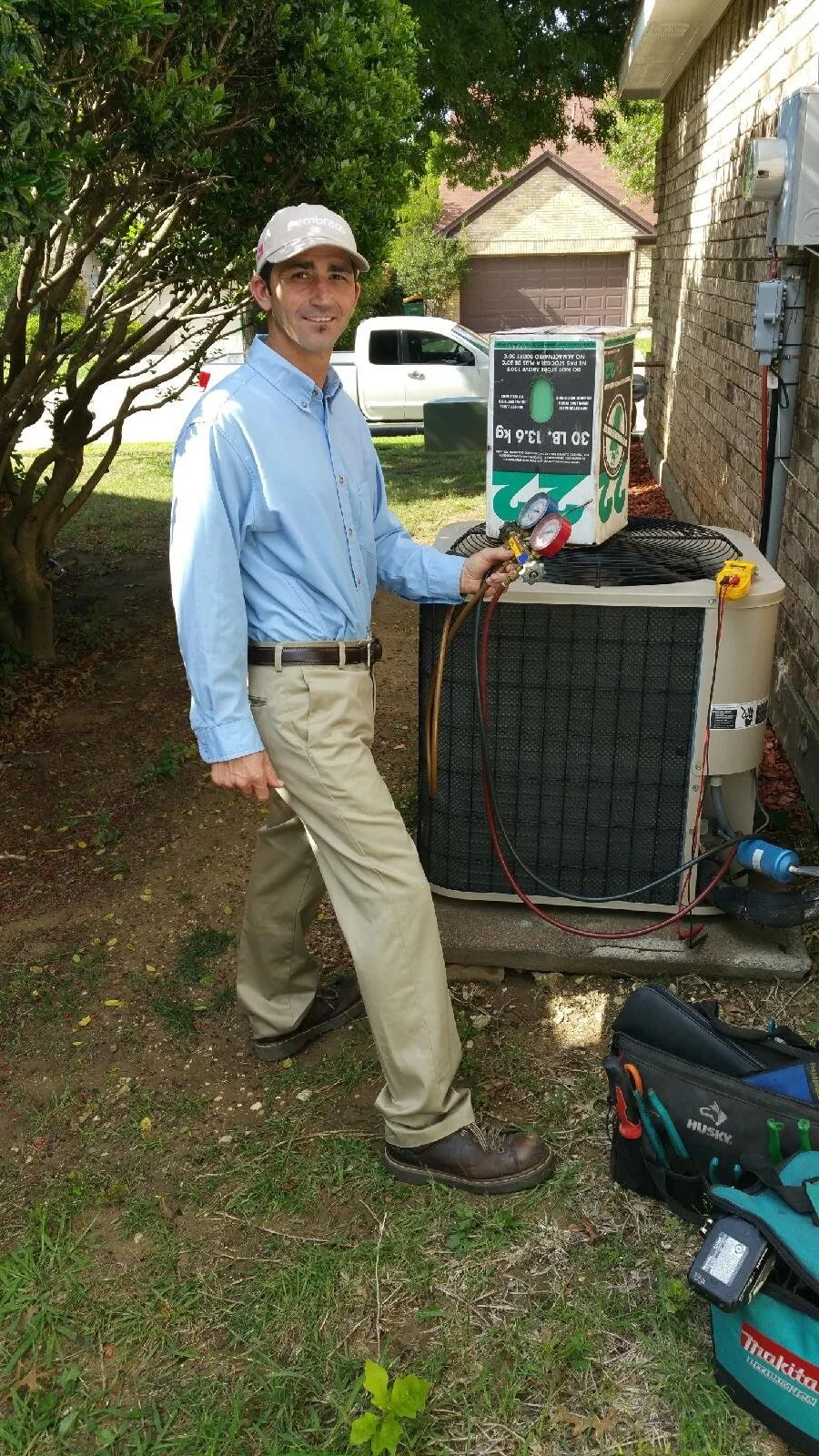 A man working on an air conditioning unit outside a house, holding HVAC gauges, wearing a light blue shirt, khaki pants, a cap, and standing on grassy ground with tools nearby.