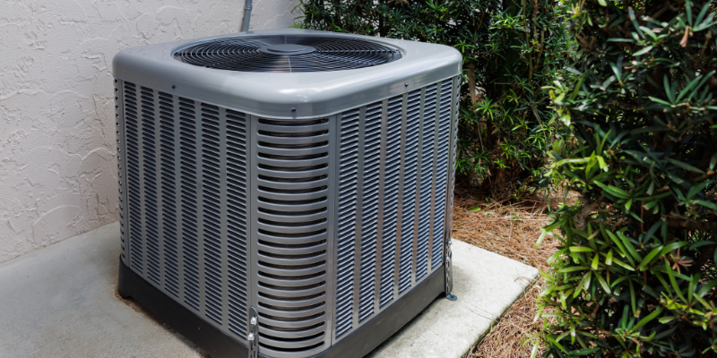 An outdoor central air conditioning unit covered with a metal grille, situated on a concrete slab next to a beige stucco wall and surrounded by green bushes.