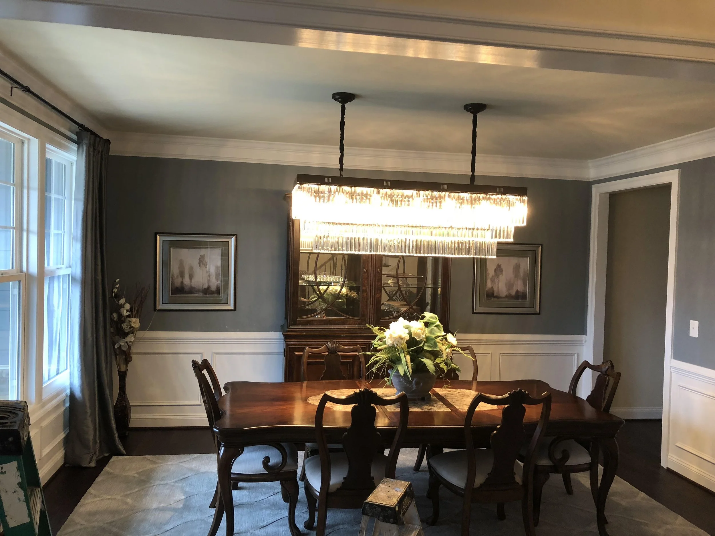 A dining room with a rectangular wooden dining table and six matching chairs. A floral centerpiece on the table. A large modern rectangular chandelier hangs above the table. There are framed pictures on the wall, a window with curtains, and a sideboard or cabinet behind the table.