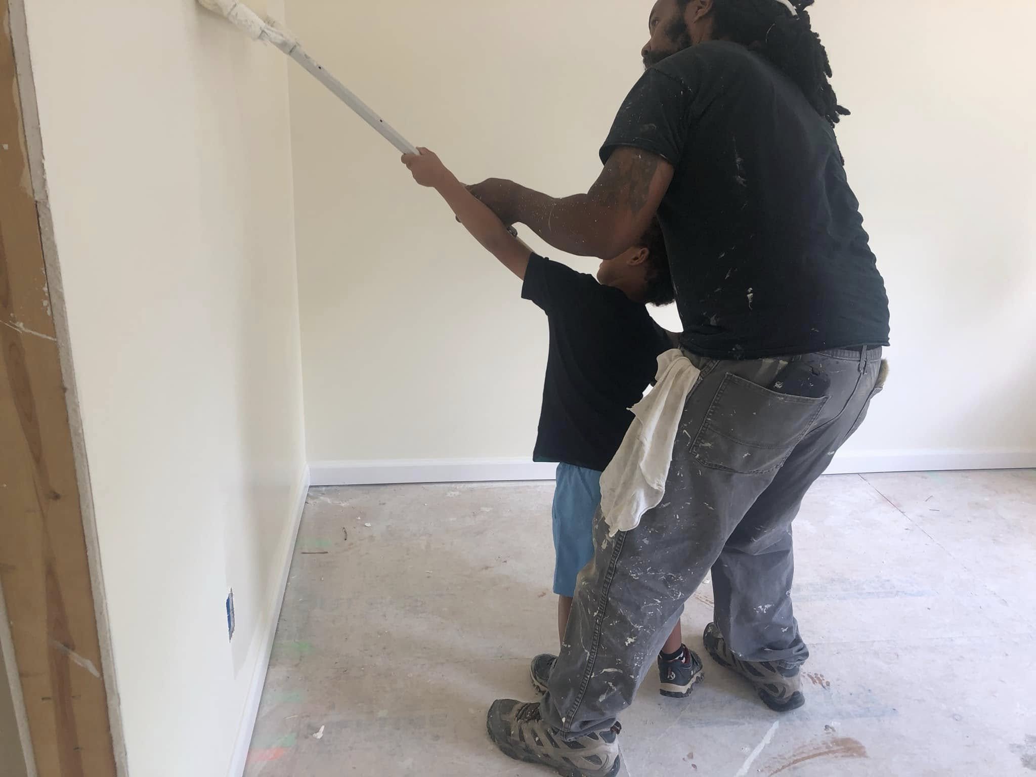 Two men painting a wall together, one standing and the other kneeling, both with paint splatters on their clothes in a room under renovation.