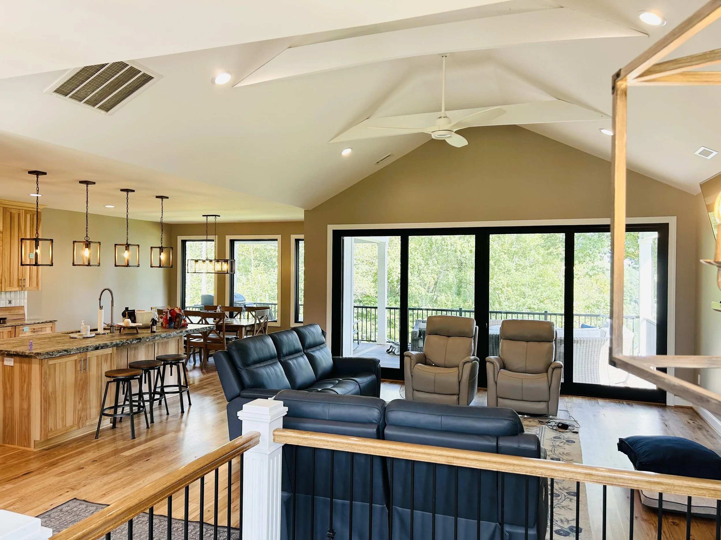 Open-concept living room and kitchen with large windows, vaulted ceiling, hanging pendant lights, ceiling fan, and a view of green outside.
