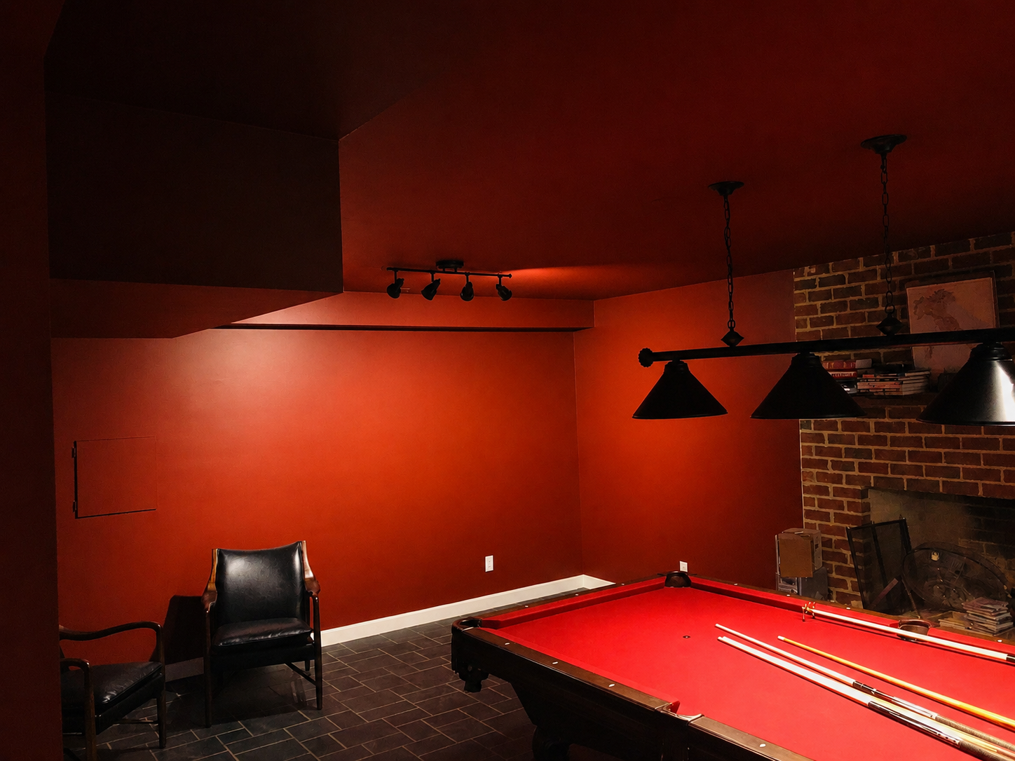 Interior of a game room featuring a red pool table with pool cues, black chairs, a brick fireplace, and dim lighting.