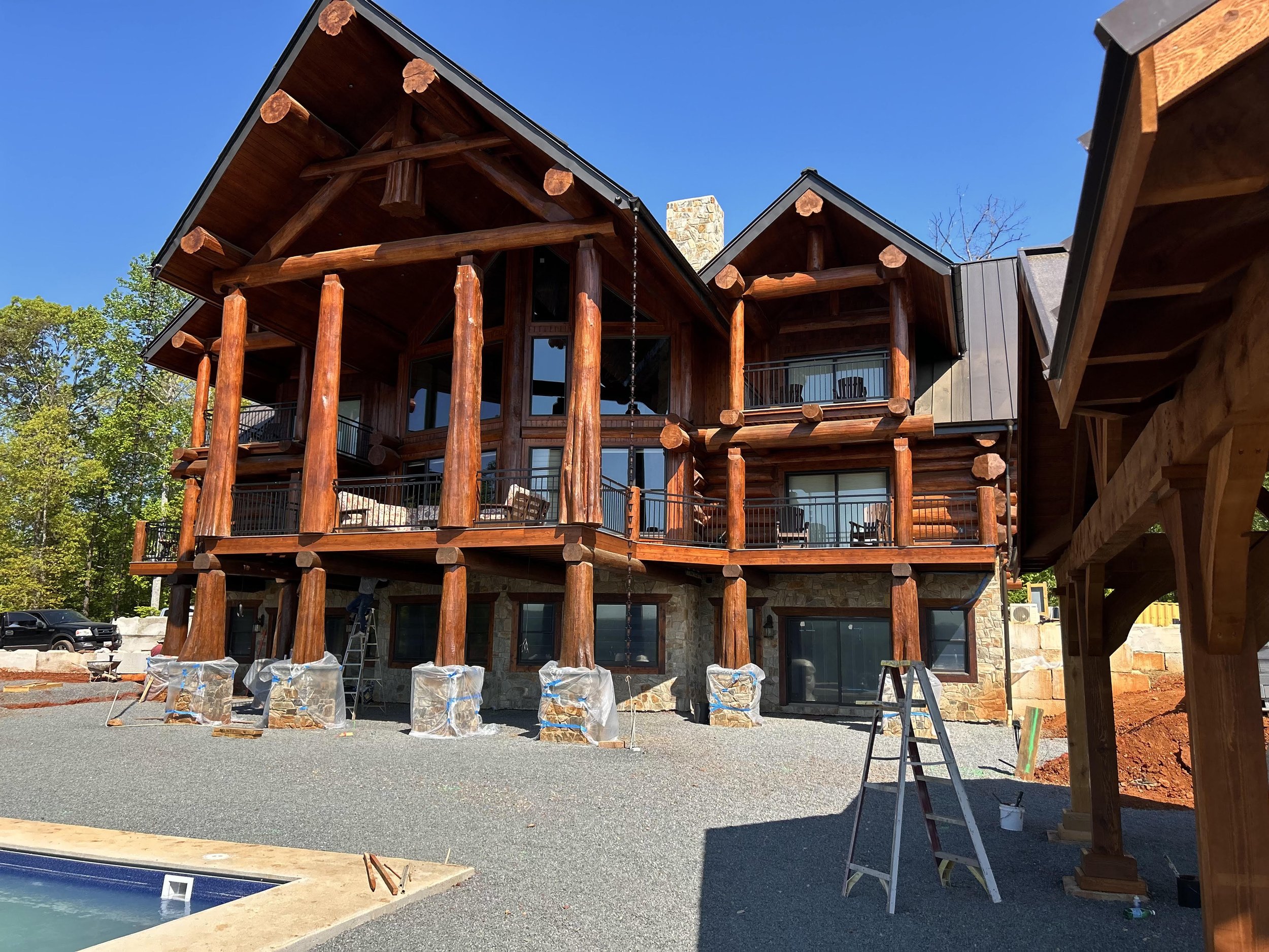 Under construction view of a multi-story log cabin house with wooden logs, large windows, and a stone foundation, surrounded by trees and a clear blue sky.