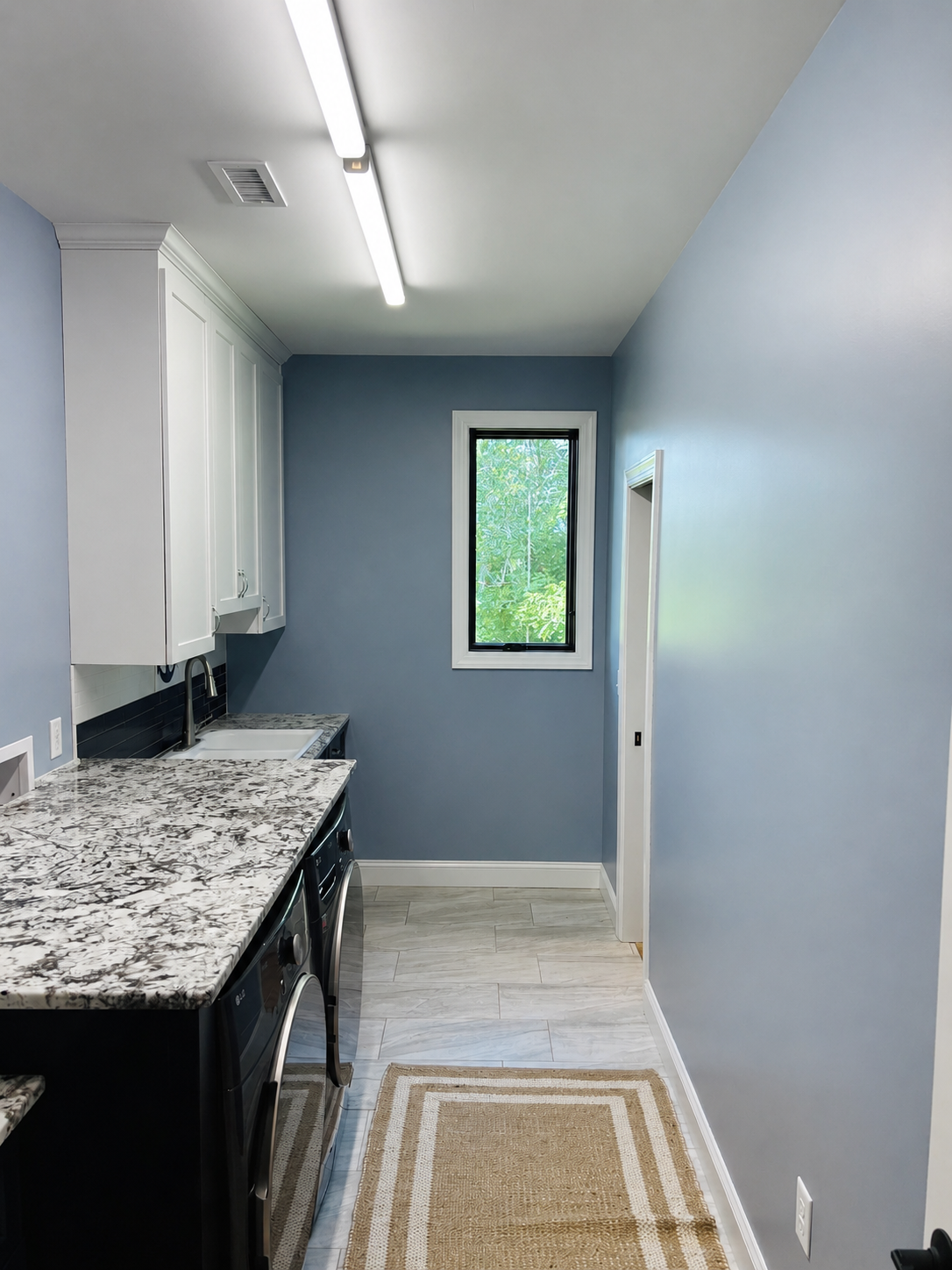 A small laundry room with blue walls, white cabinets, a granite countertop, a sink, a front-loading washing machine, a window showing trees outside, and a beige rug on the floor.
