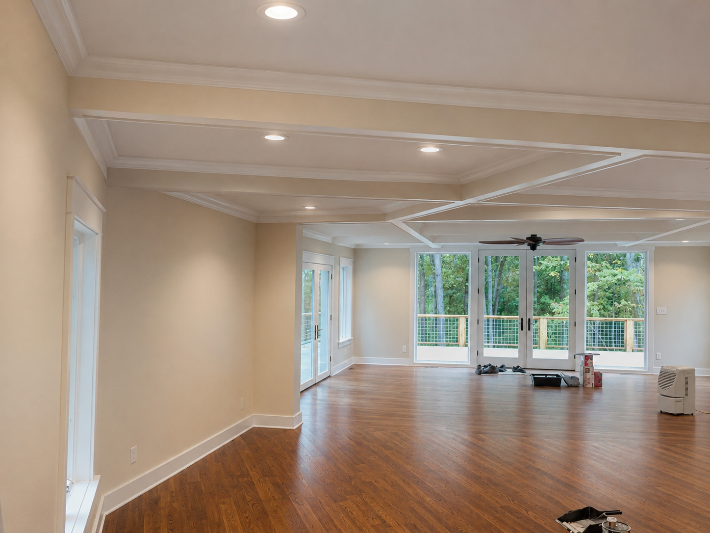 Empty living room with hardwood floors, cream-colored walls, large windows and glass doors leading to a balcony, ceiling fan, and recessed lighting.