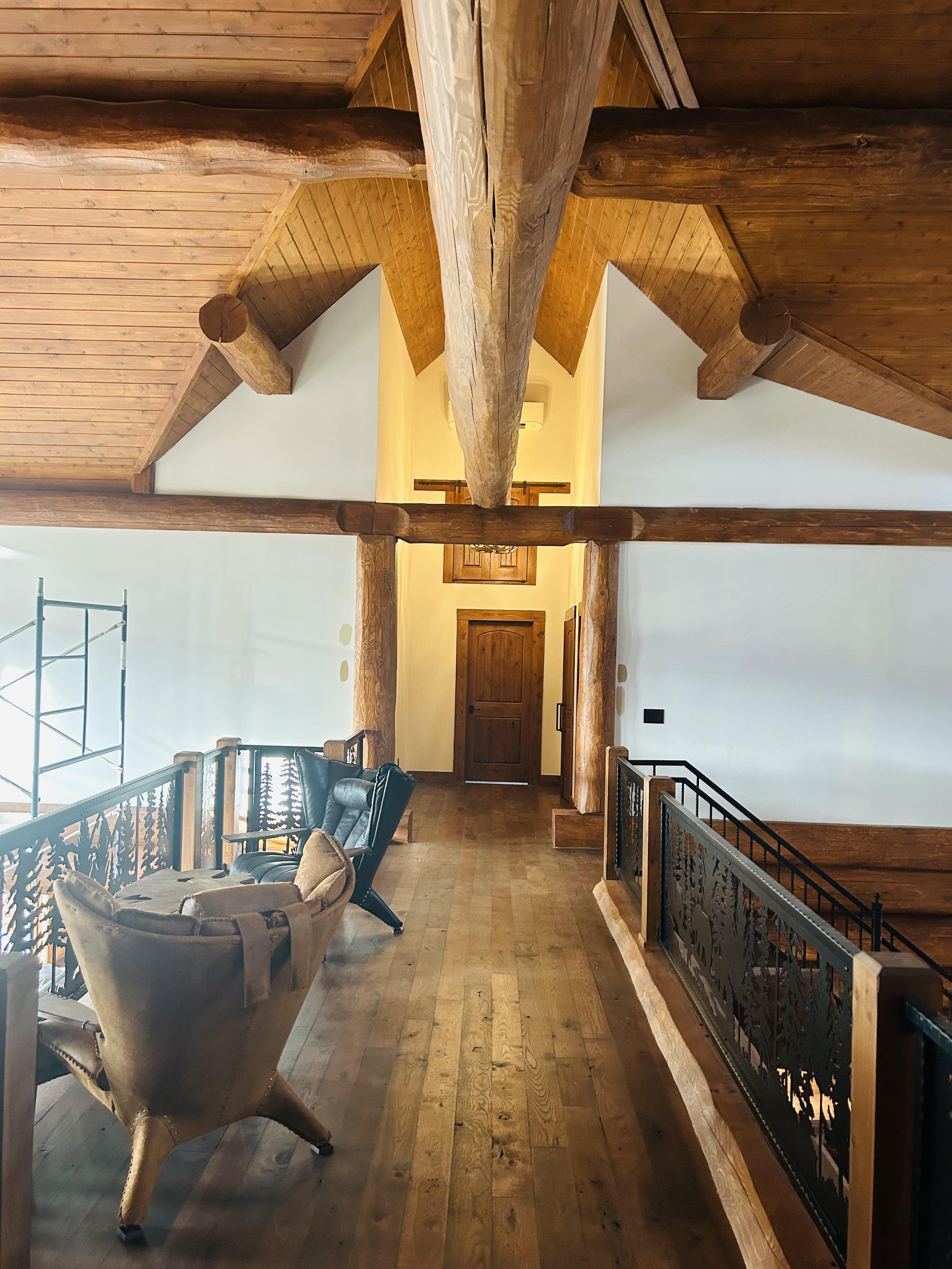 Interior of a wooden house with a high ceiling, wooden beams, and a staircase with a black wrought-iron railing. There is a seating area with a leather armchair and a beige upholstered armchair. The walls are painted white, and there is a wooden door at the end of the hallway.