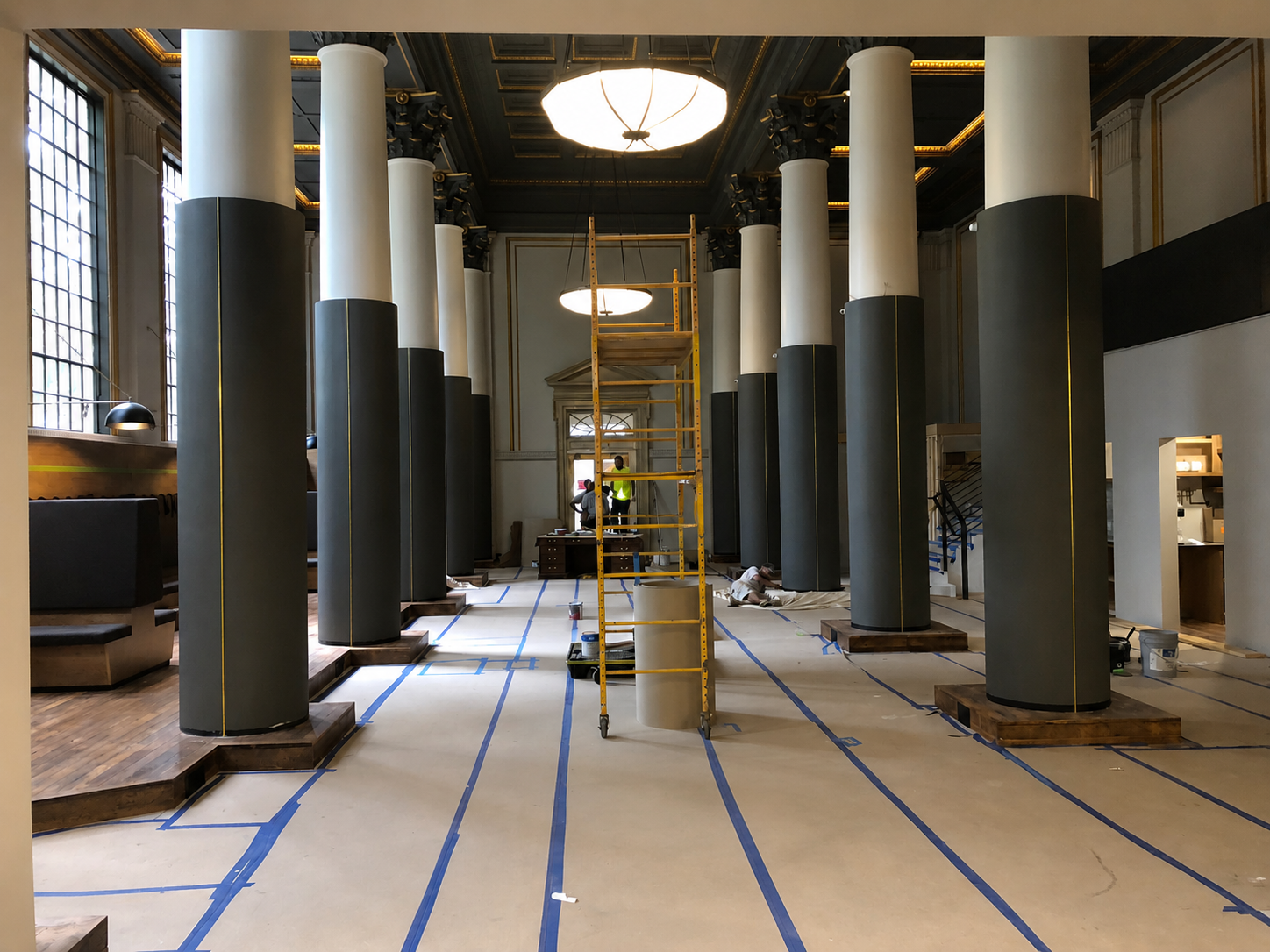 Interior of a historic building under renovation with tall white columns, large windows, and construction workers building or repairing. Blue tape lines on the floor mark areas for work, and a yellow scaffold is positioned in the center.