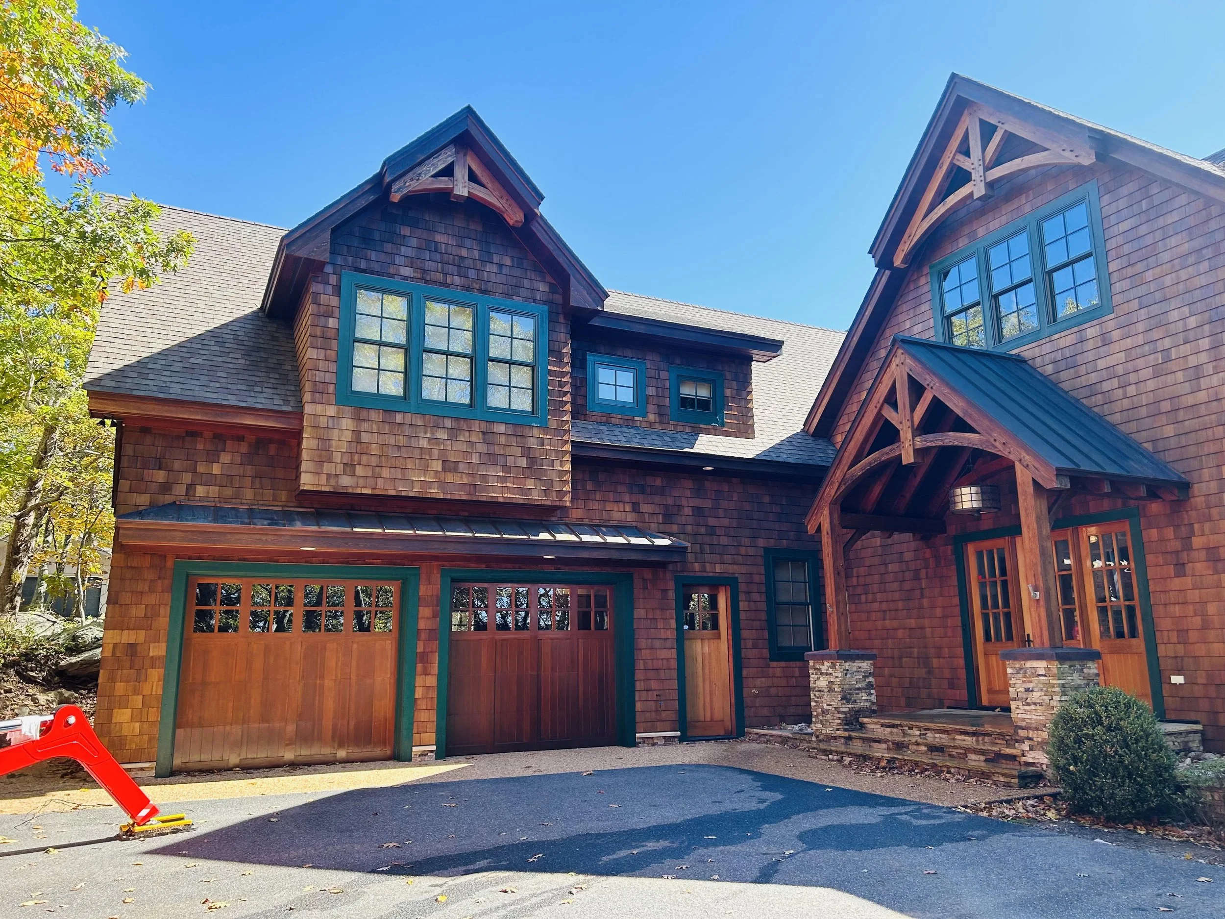 A house with wooden shingles, multiple large windows with teal frames, and a steep roof with decorative wooden beams. There is a driveway in front with a red and yellow toy on it, and trees with autumn leaves surrounding the house.