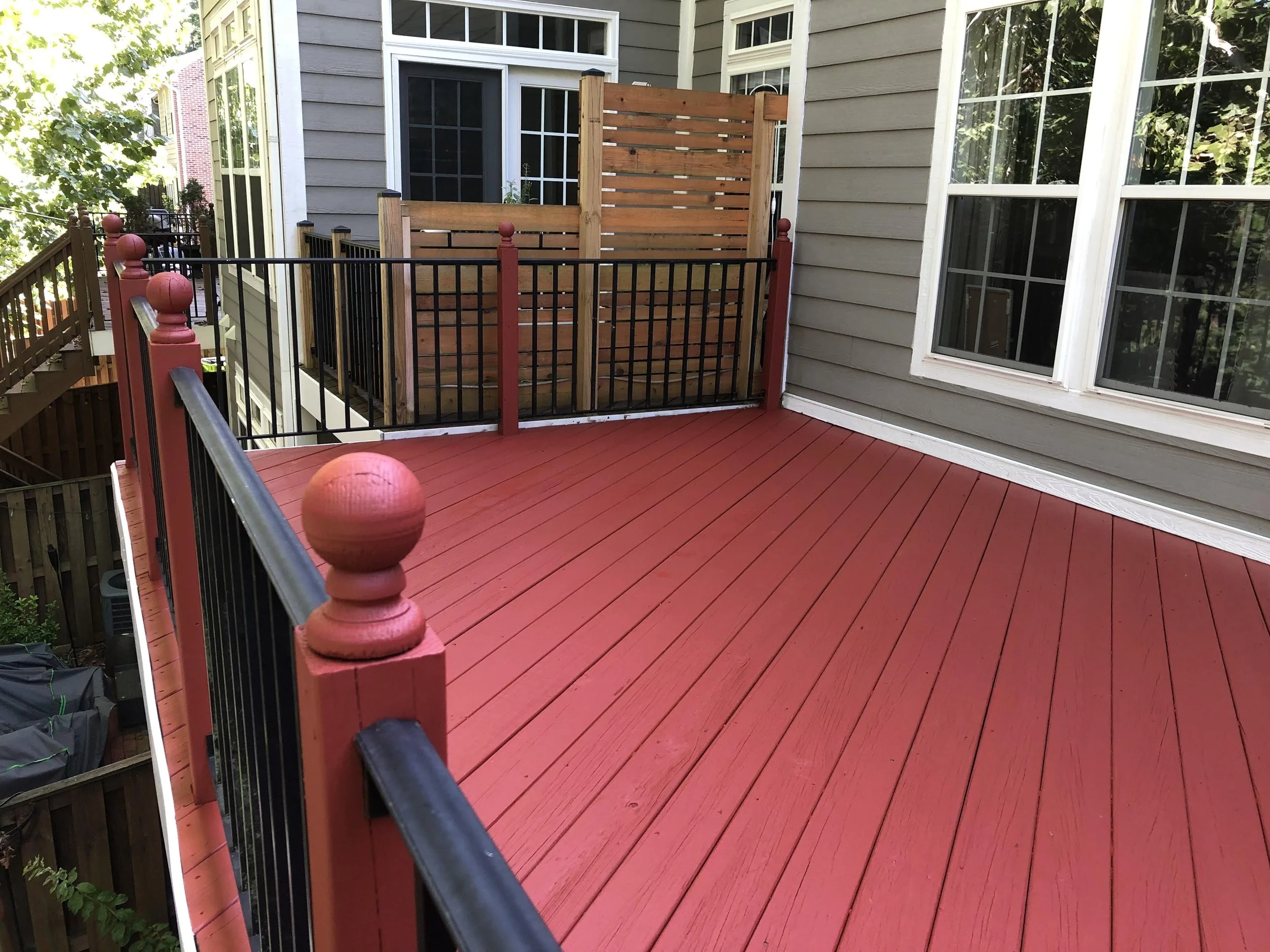 Backyard deck with red-painted wooden floorboards, a black metal railing with red wooden posts, and a privacy fence made of horizontal wooden slats. The house features gray siding and large white-framed windows, with trees and greenery reflected in t