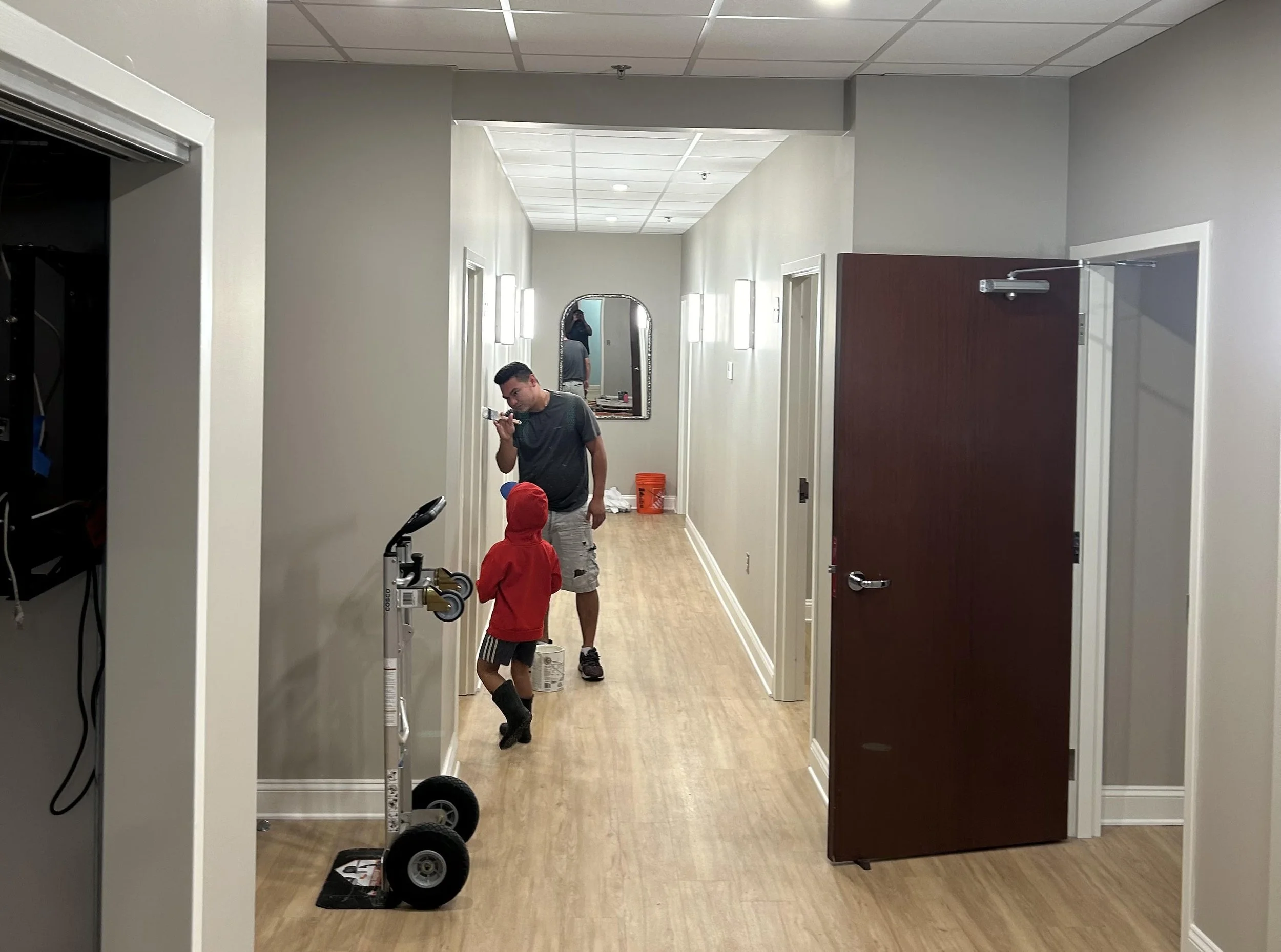 A hallway with white walls and light wood flooring. Two men and a child are in the corridor, with one man holding a toothbrush. There is a mirror on the wall reflecting another person and the ceiling has fluorescent lights. A black equipment cart is visible on the left, and an orange bucket is on the floor in the background.