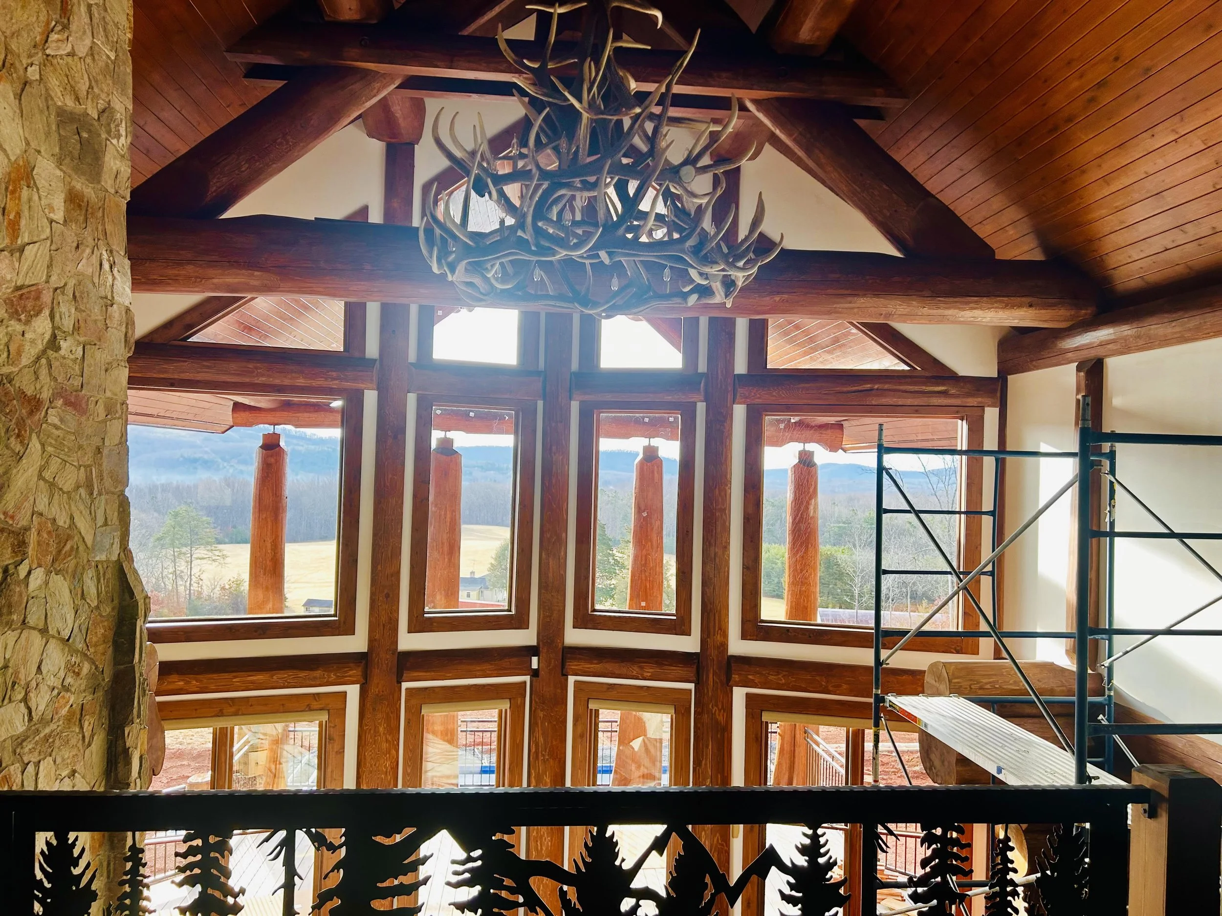 Interior of a house under construction with large wooden beams, windows, and a chandelier made of antlers hanging from the ceiling.