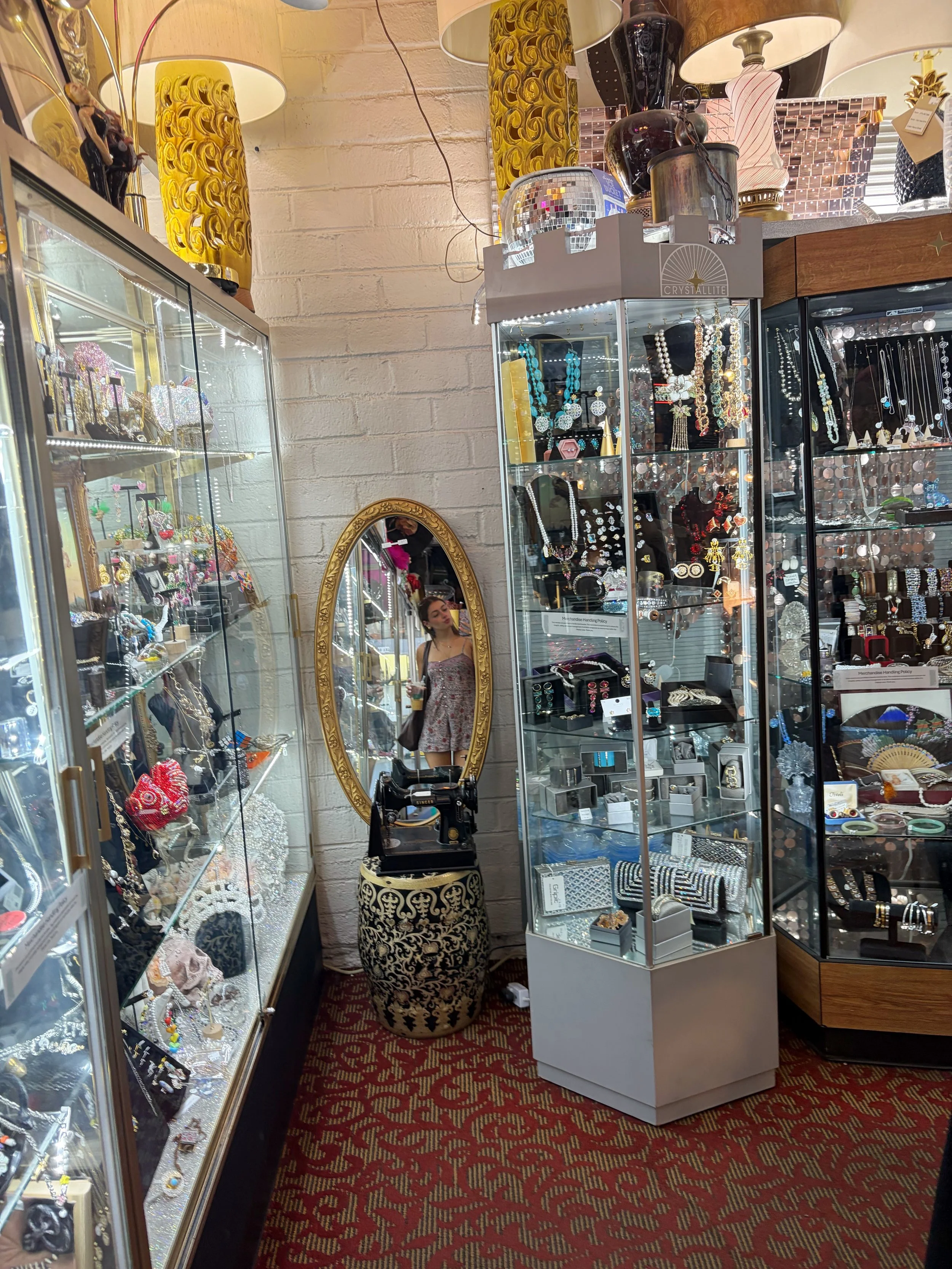 Interior de loja de antiguidades e bijuterias, com estantes de vidro exibindo joias, acessórios e objetos decorativos, espelho oval dourado, tapete vermelho com padrão dourado, e diversas luminárias e vasos decorativos no fundo.