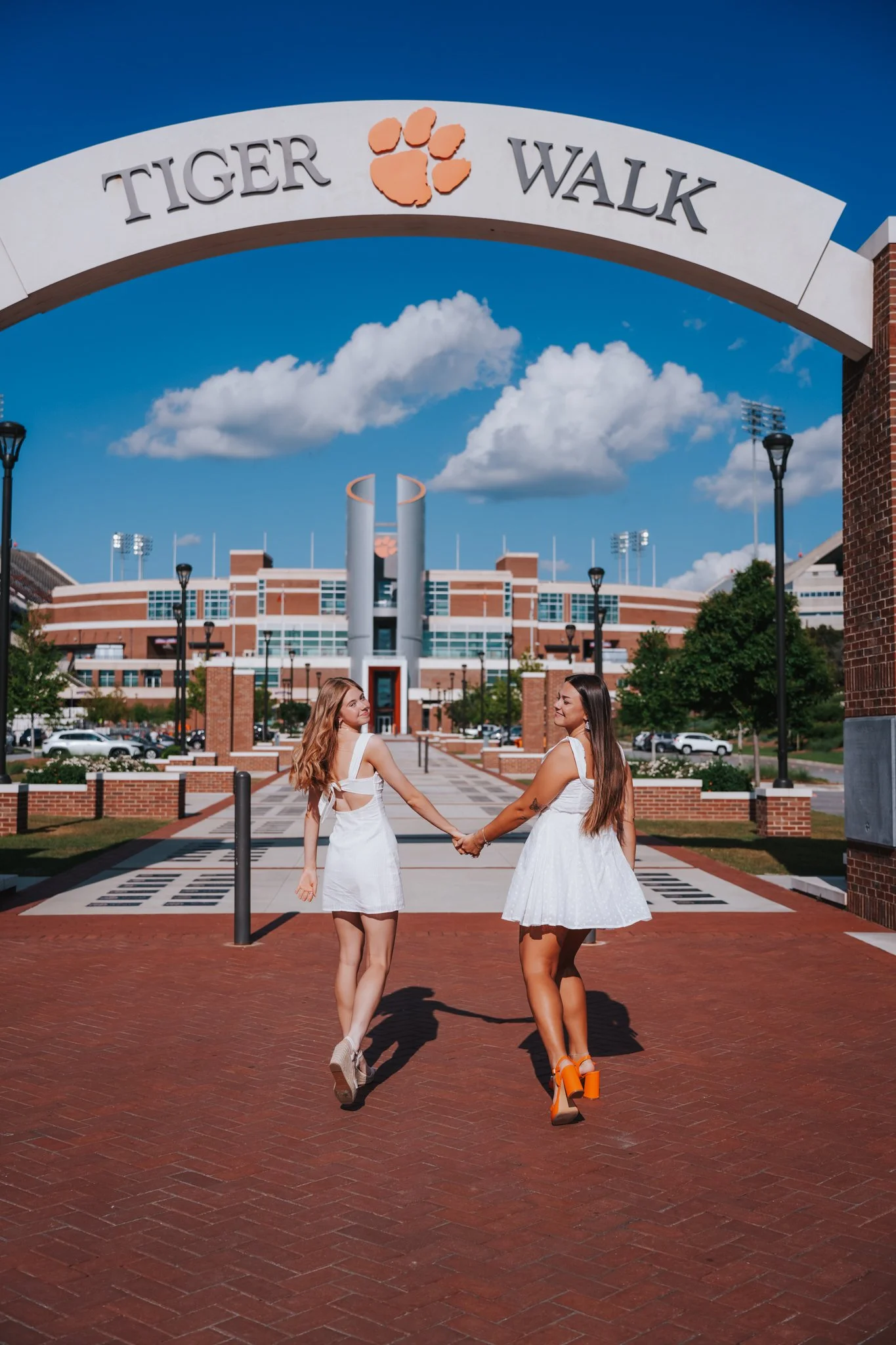 Duas mulheres jovens, de vestidos brancos, se segurando pela mão na frente de uma entrada de estádio chamada Tiger Walk, sob um céu azul com nuvens brancas.