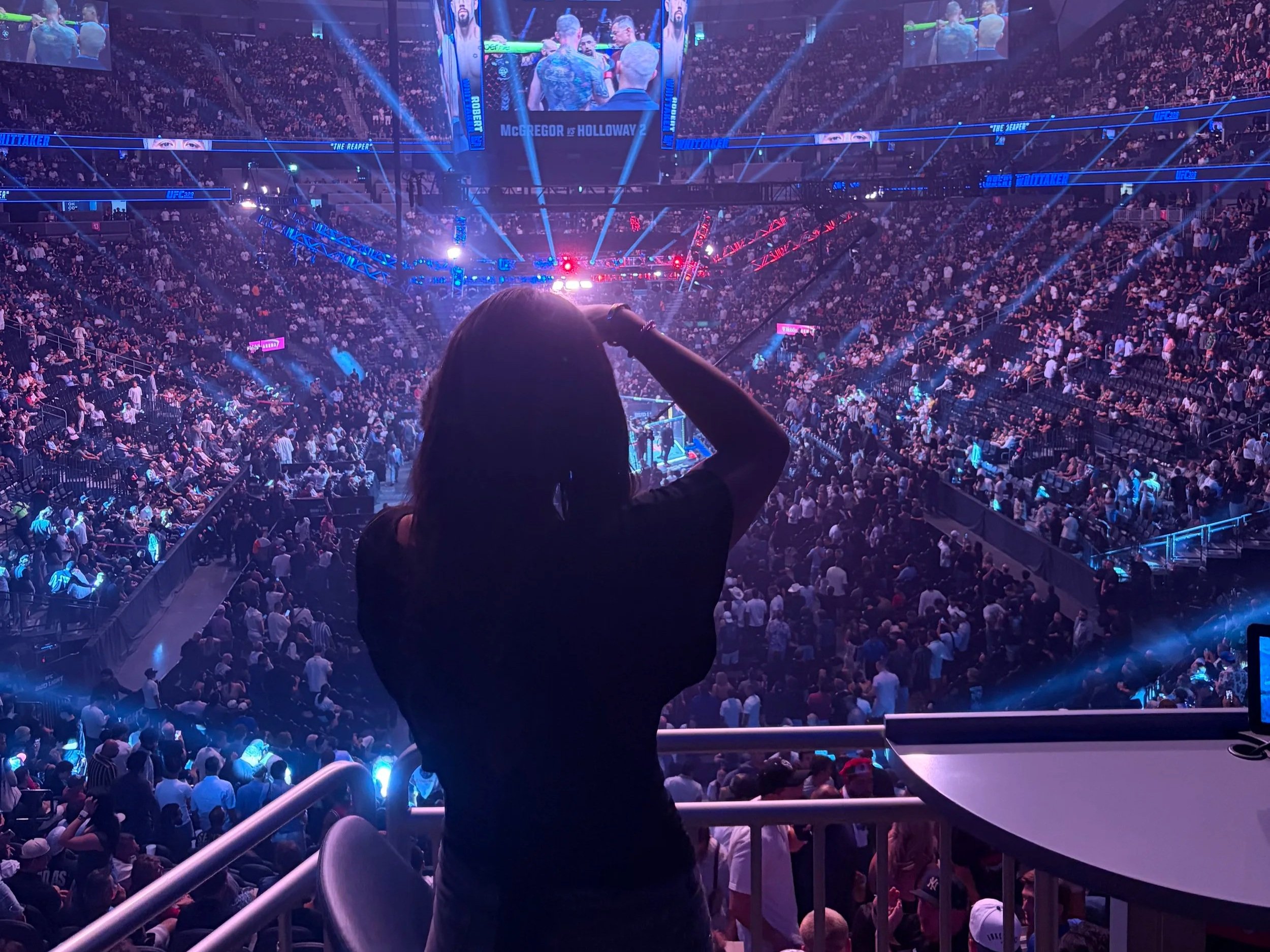 Pessoa assistindo a uma luta de MMA no ringue, de costas, com uma grande audiência em um arena iluminada com luzes de neón.
