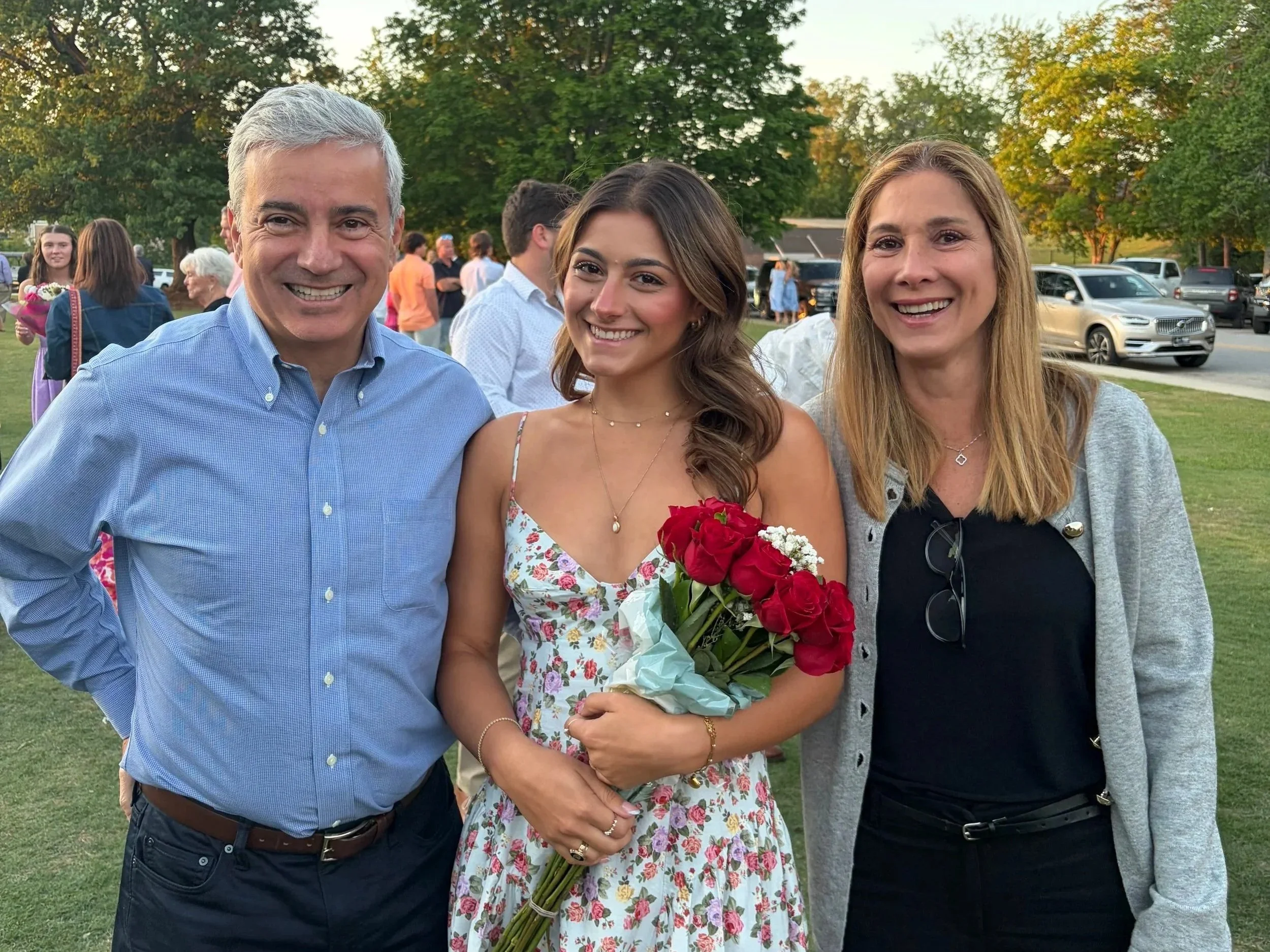 Três pessoas sorridentes posando ao ar livre, uma delas segurando um buquê de rosas vermelhas.