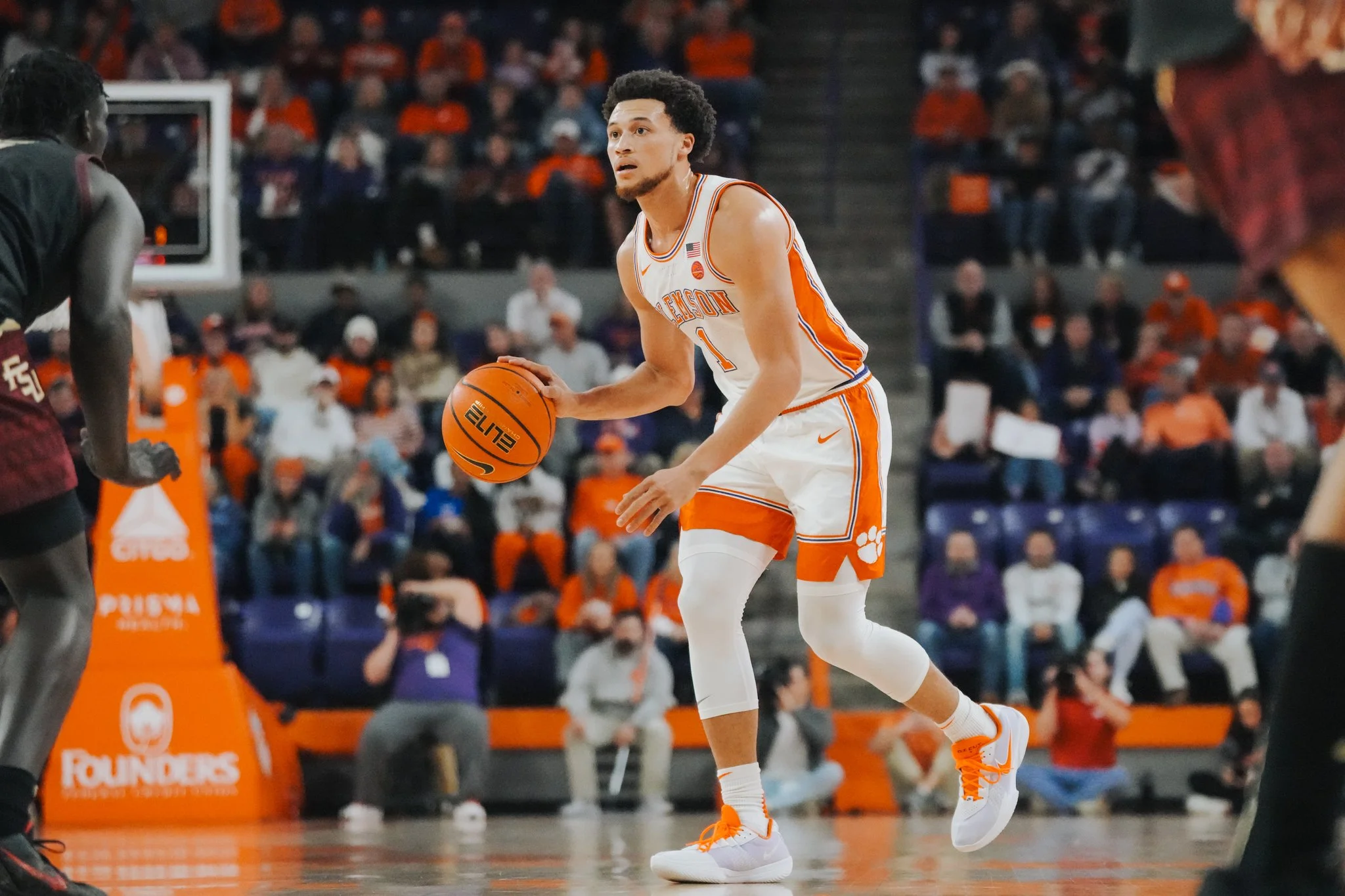 Jogador de basquete com chuteira branca e uniforme laranja e branco, segurando uma bola de basquete no chão, em quadra com público ao fundo.