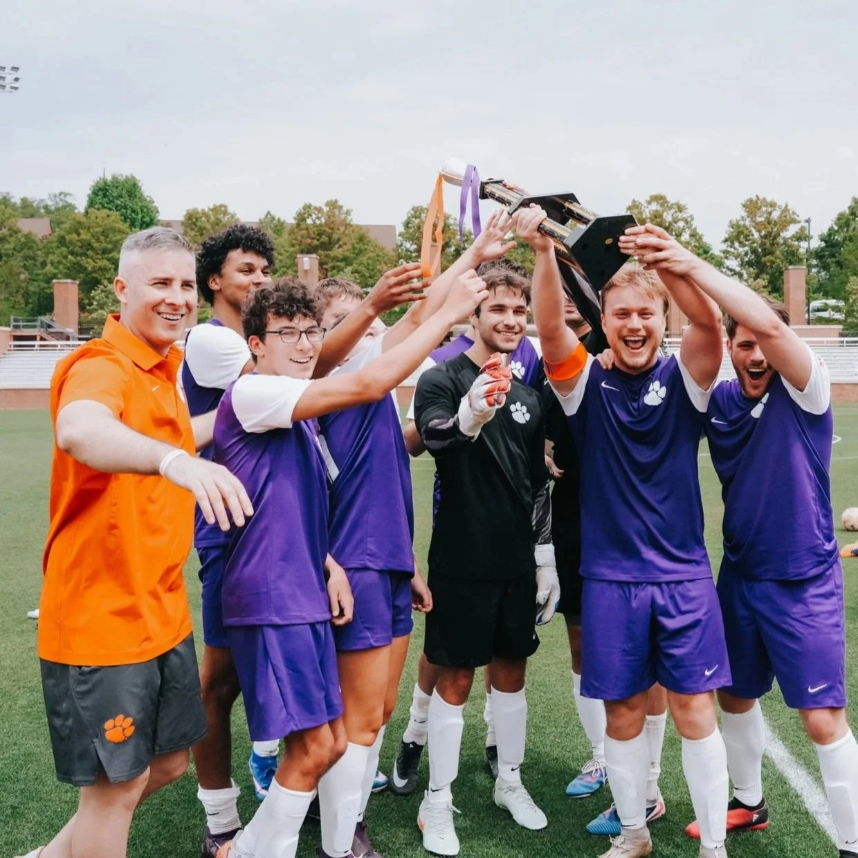 Time de futebol celebrando uma vitória com troféu, com jogadores usando uniformes roxos e um treinador com camisa laranja, em um campo ao ar livre.