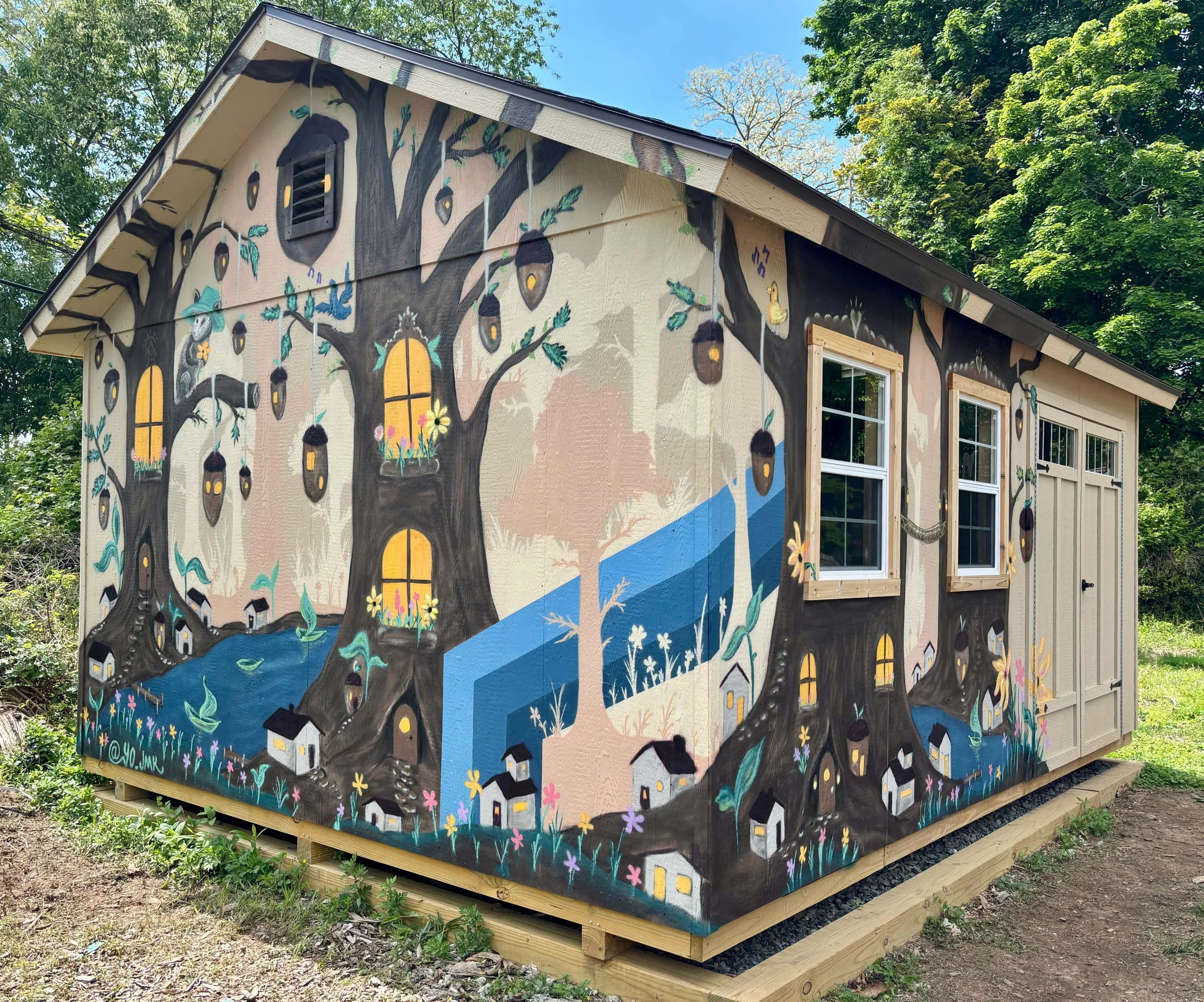 A small shed with a mural of a large tree with birdhouses, flowers, and a window painted on its side, surrounded by trees and greenery.