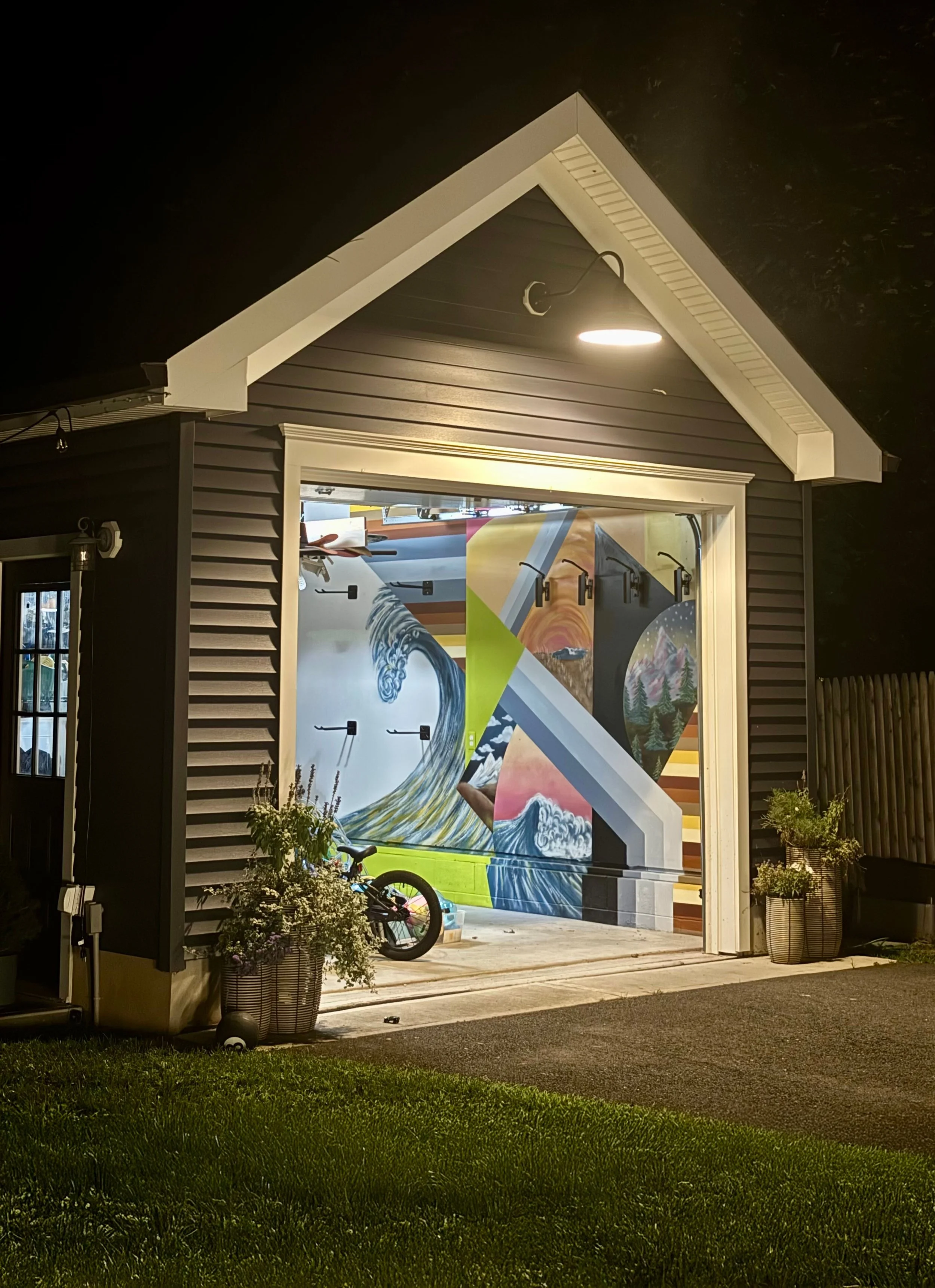 Night view of a house with a dark exterior, featuring a large window displaying colorful artwork with mountain and ocean scenes, and a bicycle outside on the sidewalk.