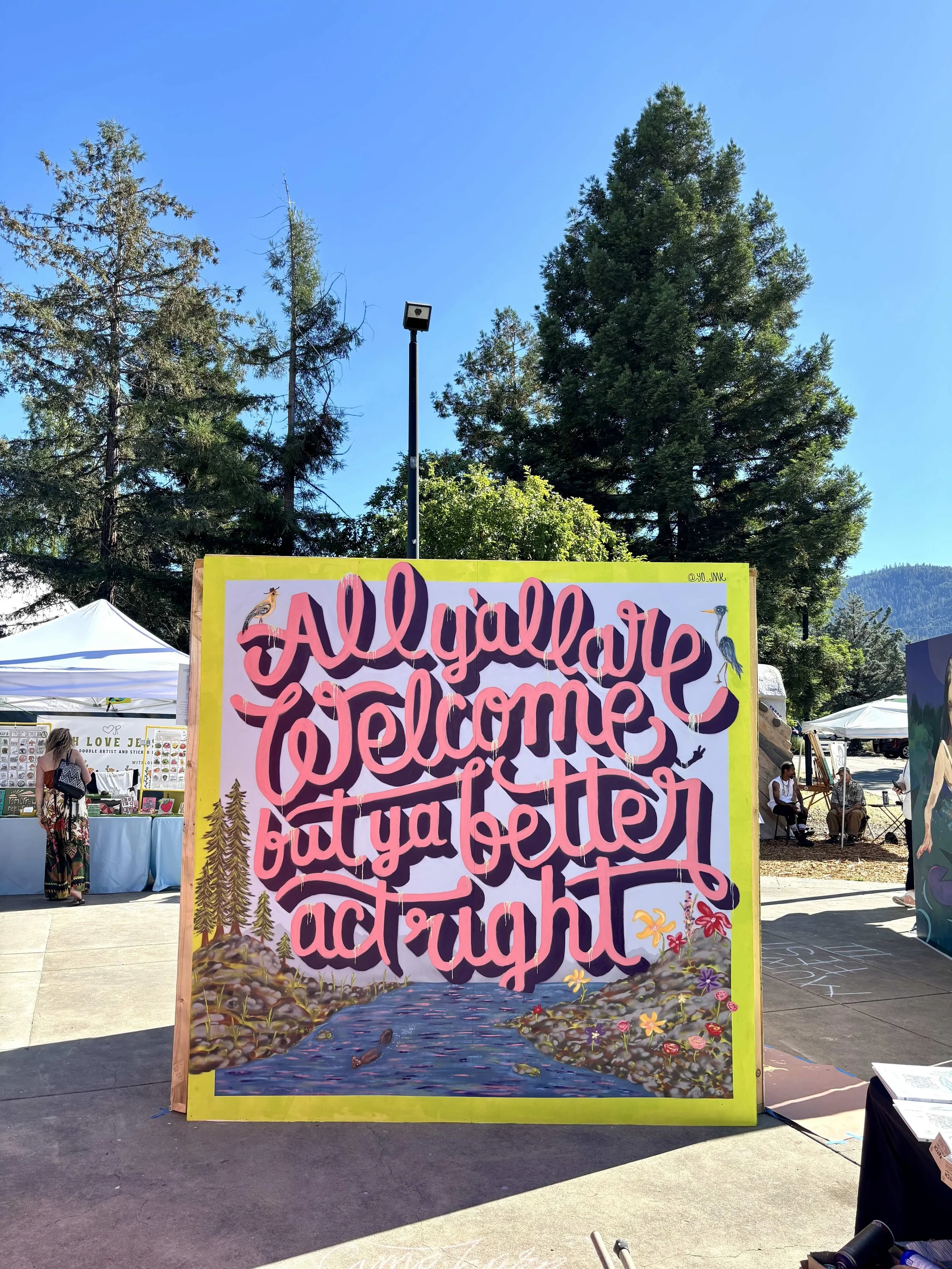 A colorful sign with hand-lettered text that says 'Allergy Welcome', with illustrations of trees, flowers, a river, and birds, at an outdoor event with tents and trees in the background.