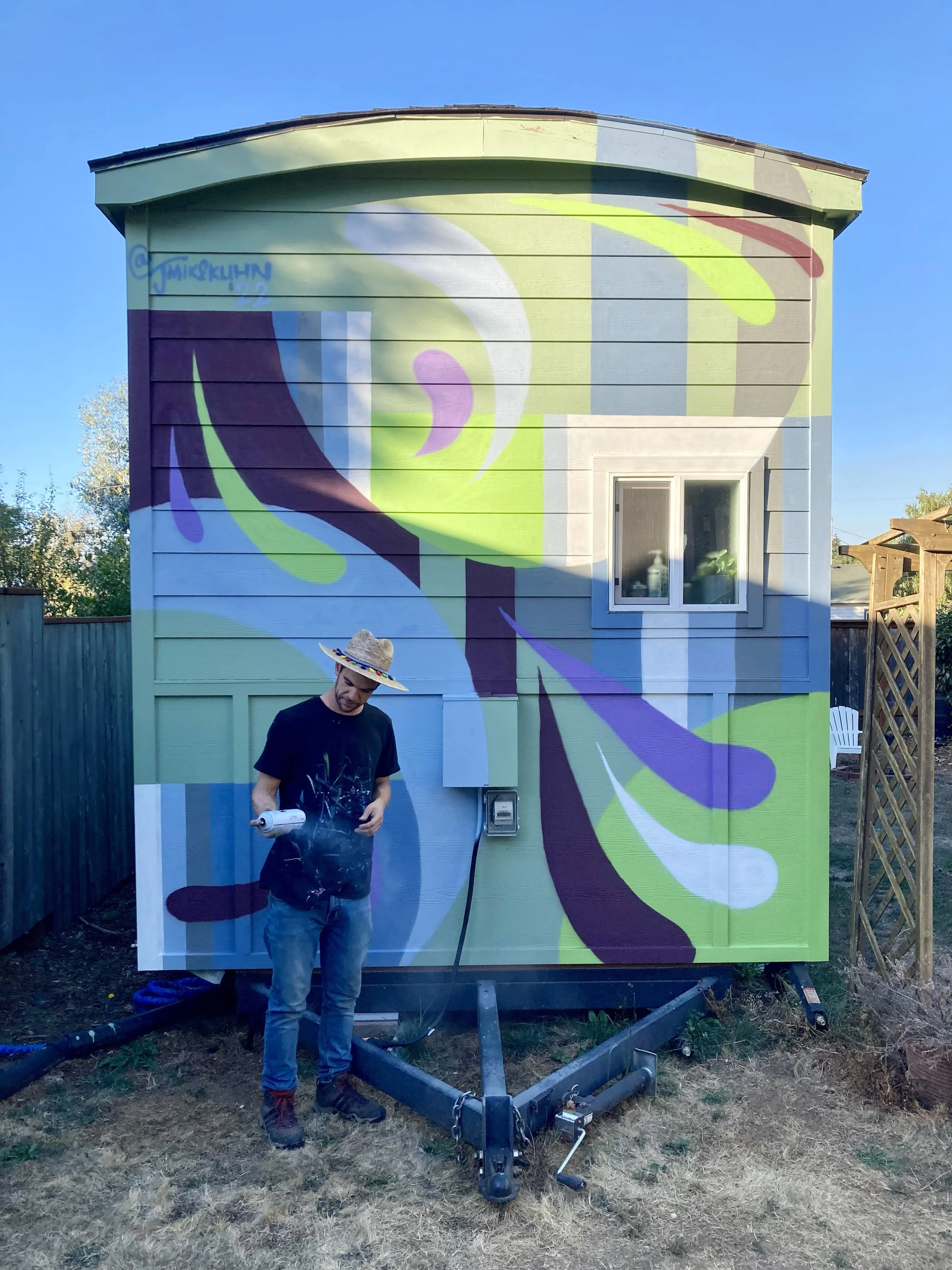 A man standing in front of a colorful mural on the side of a house, holding a spray paint can. The mural features abstract shapes with vibrant colors including purple, green, yellow, and white. The man is wearing a sombrero hat and casual clothes, wi